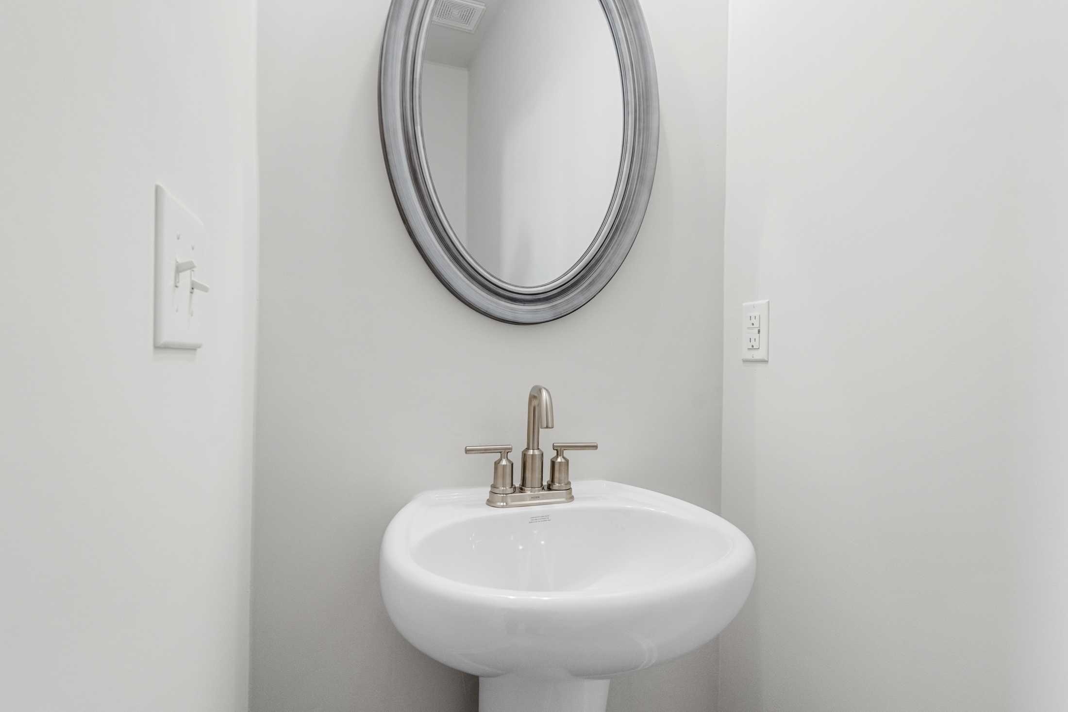 White pedestal sink with chrome faucet and round silver-framed mirror in The Glenwood A powder room