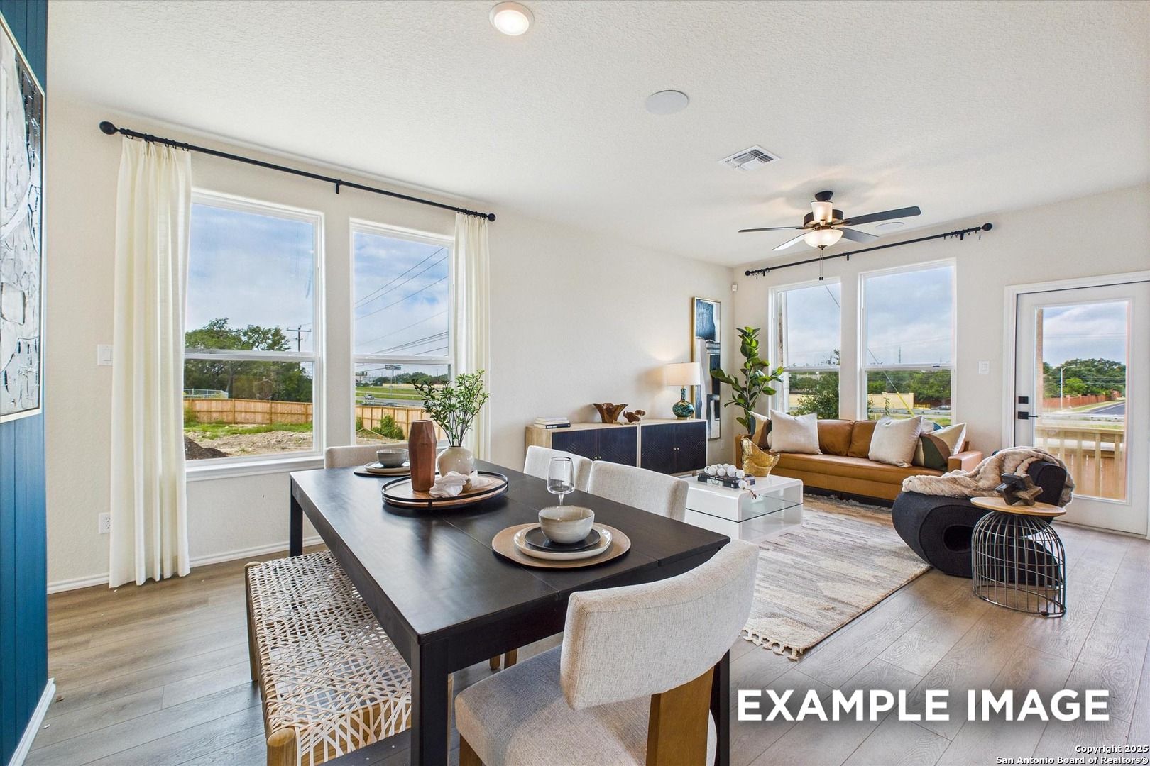 Bright open-concept living dining room with wooden floors, ceiling fan, modern sofa, and large windows in The Florence C, San Antonio