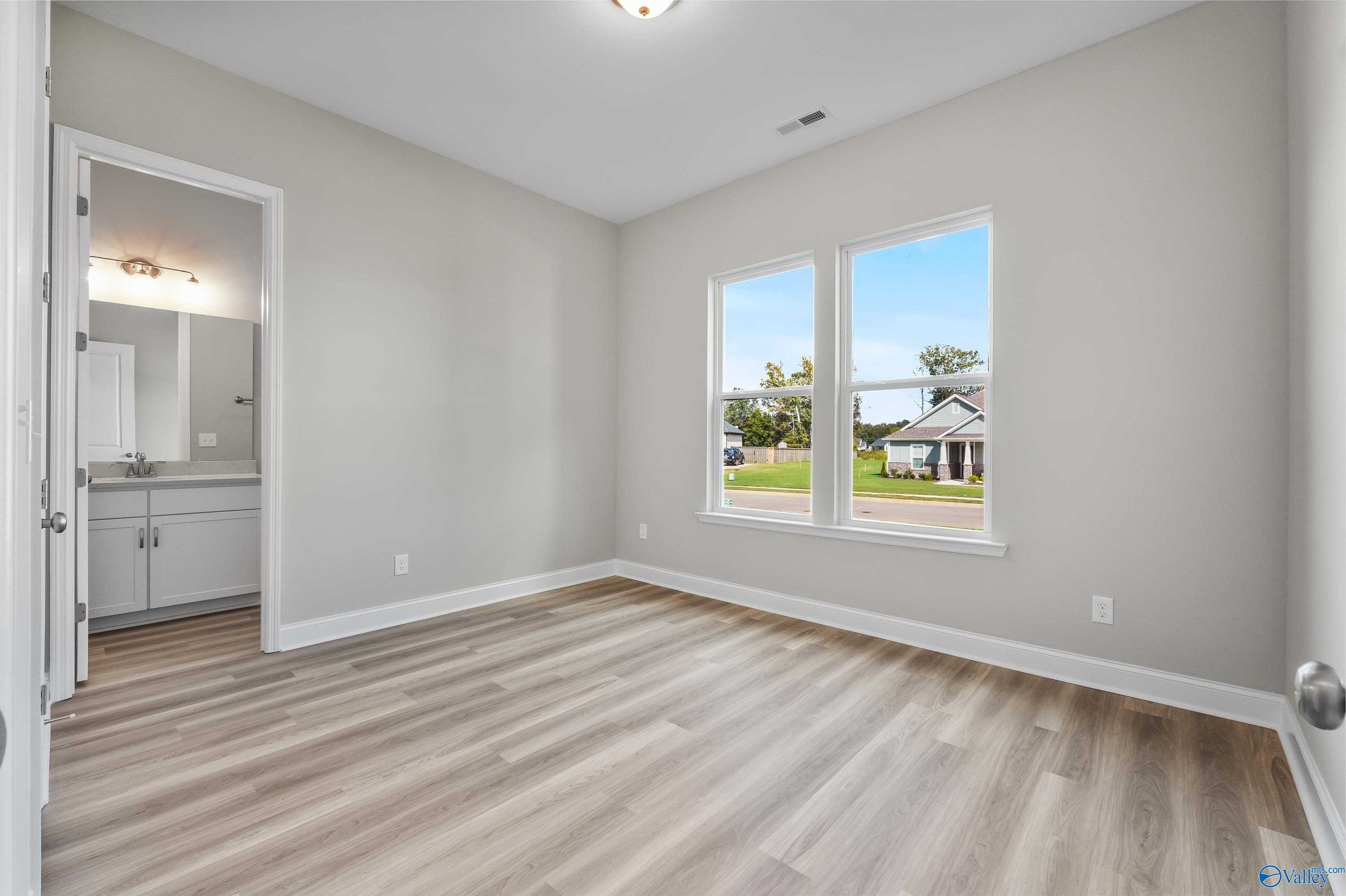 Bright secondary bedroom with en-suite bath, light gray walls, LVP flooring, and large windows overlooking green space in Evermore Homes The Haven, Owens Cross Roads, AL