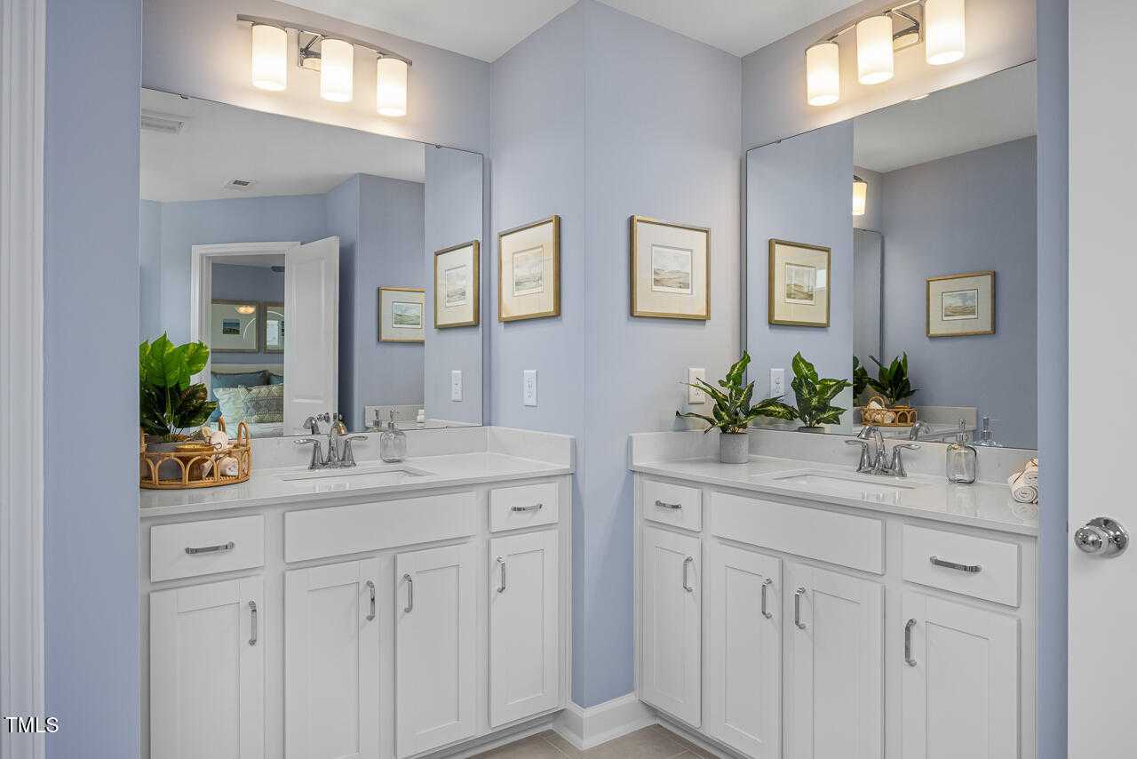 Elegant double vanity bathroom with light blue walls, white cabinets, mirrors, and potted plants in Davidson Homes The Graham Exterior, Fuquay-Varina, NC