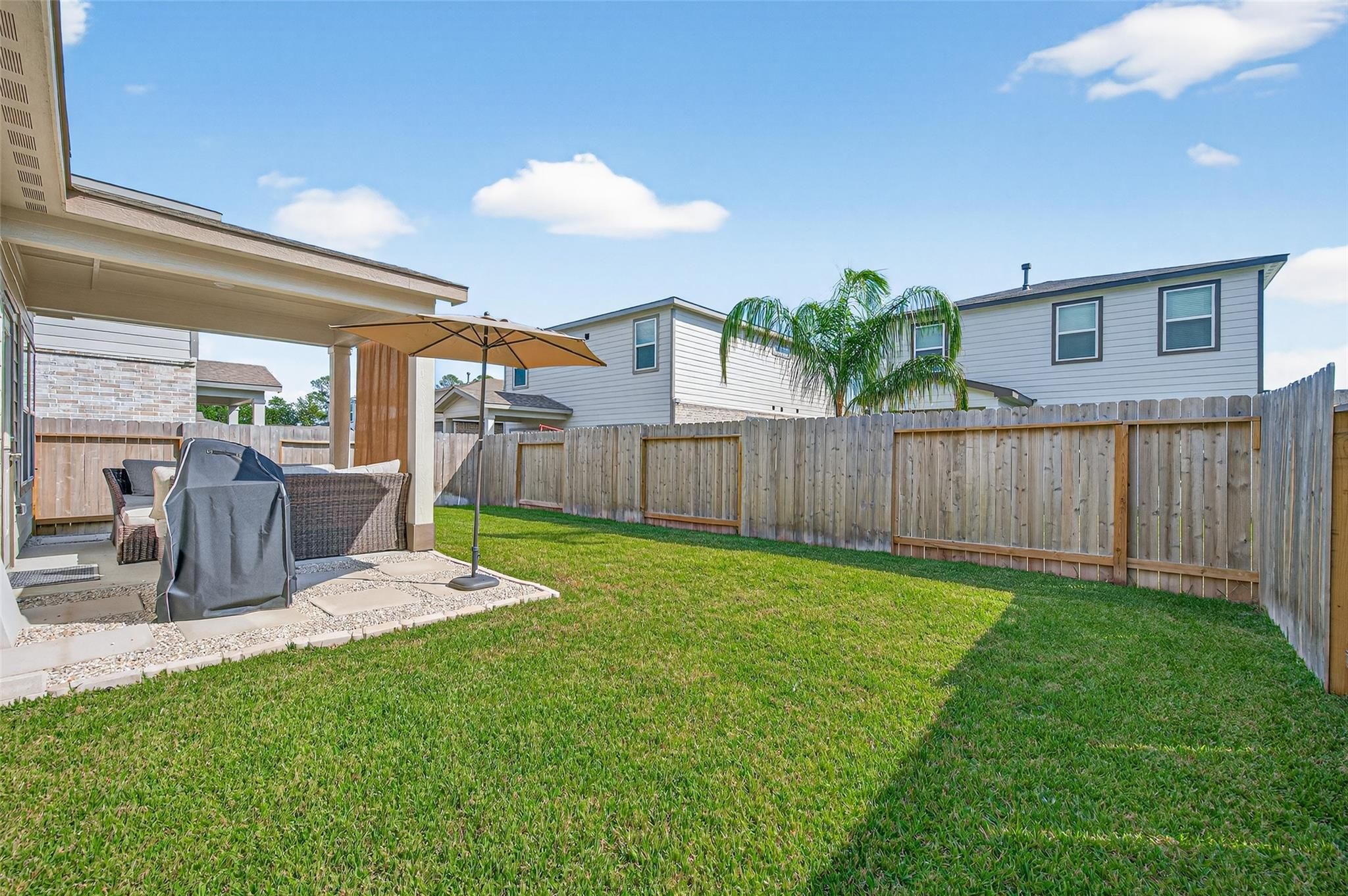 Covered back patio with shaded umbrella, covered seating, and grill in lush backyard of 5-bed Davidson Homes The Brazos E, Magnolia, TX