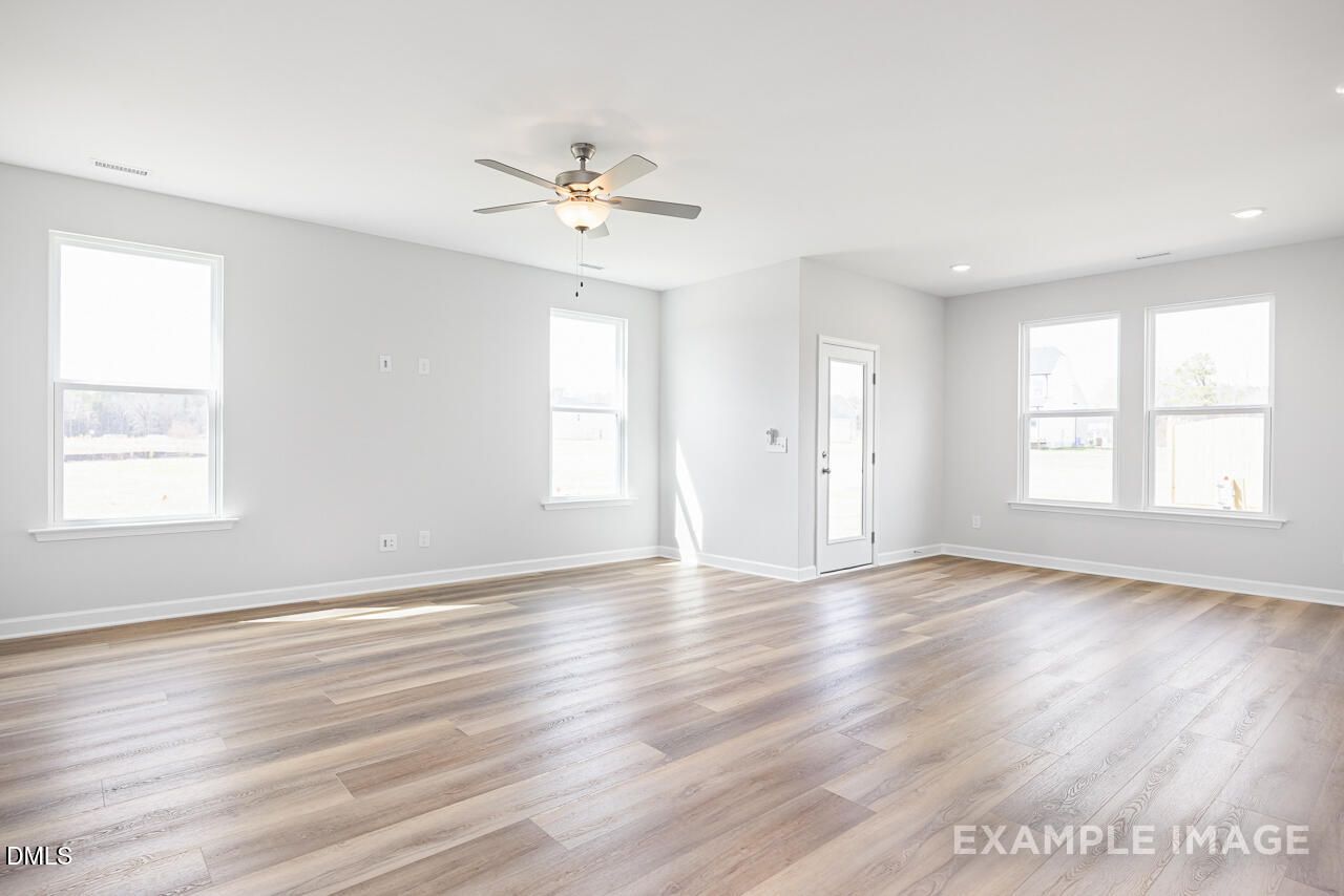 Spacious empty living room with light gray walls, hardwood floors, ceiling fan, and large sunny windows in The Daphne C home, Lillington, NC