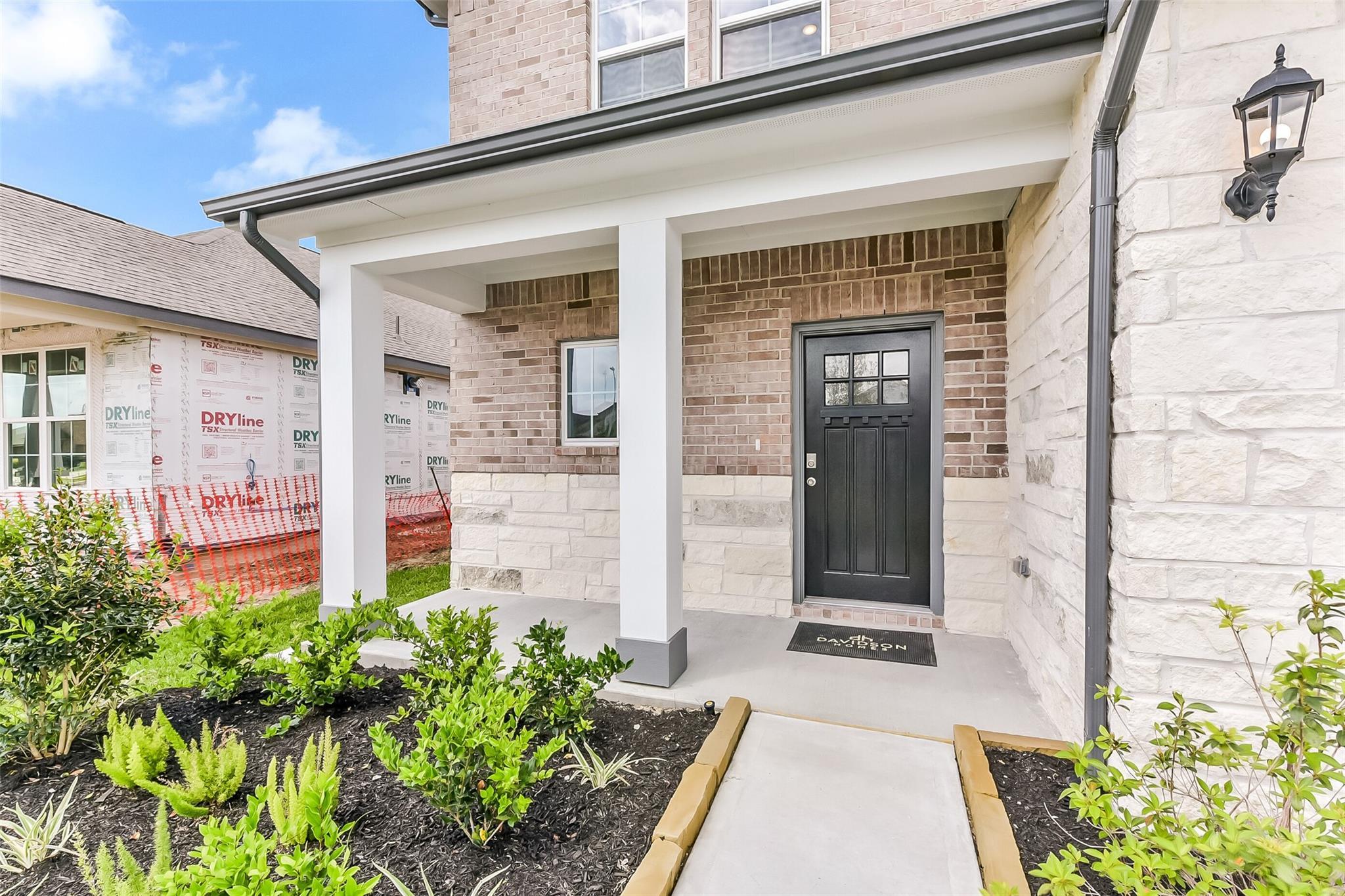 Brick and stone facade with covered front porch, black paneled door, lanterns, and lush landscaping of Davidson Homes The Tierra C in Dayton, Texas
