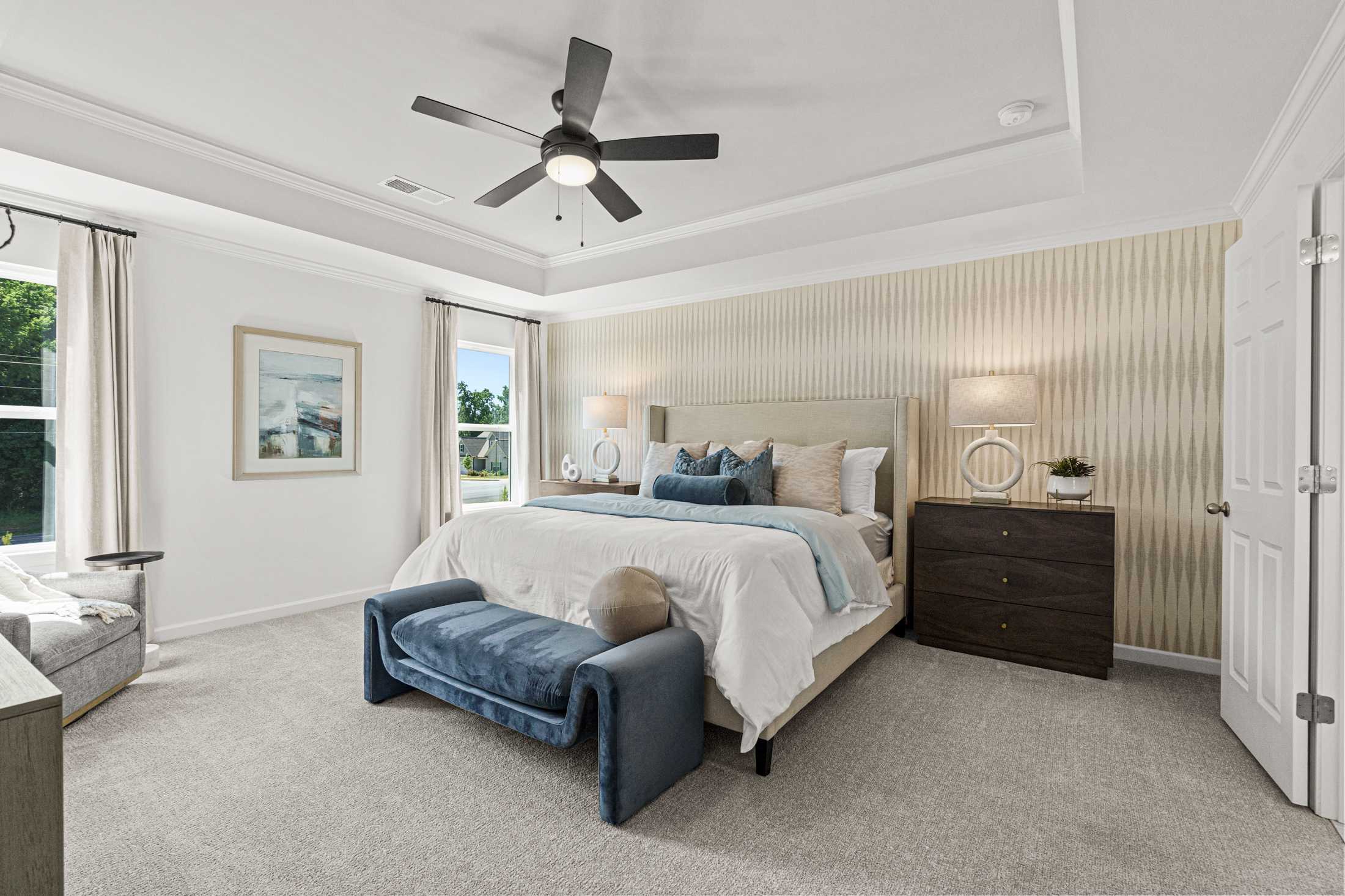 Spacious master bedroom in The Hickory A featuring king bed with blue accents, ceiling fan, and beige striped walls in Hoschton GA home