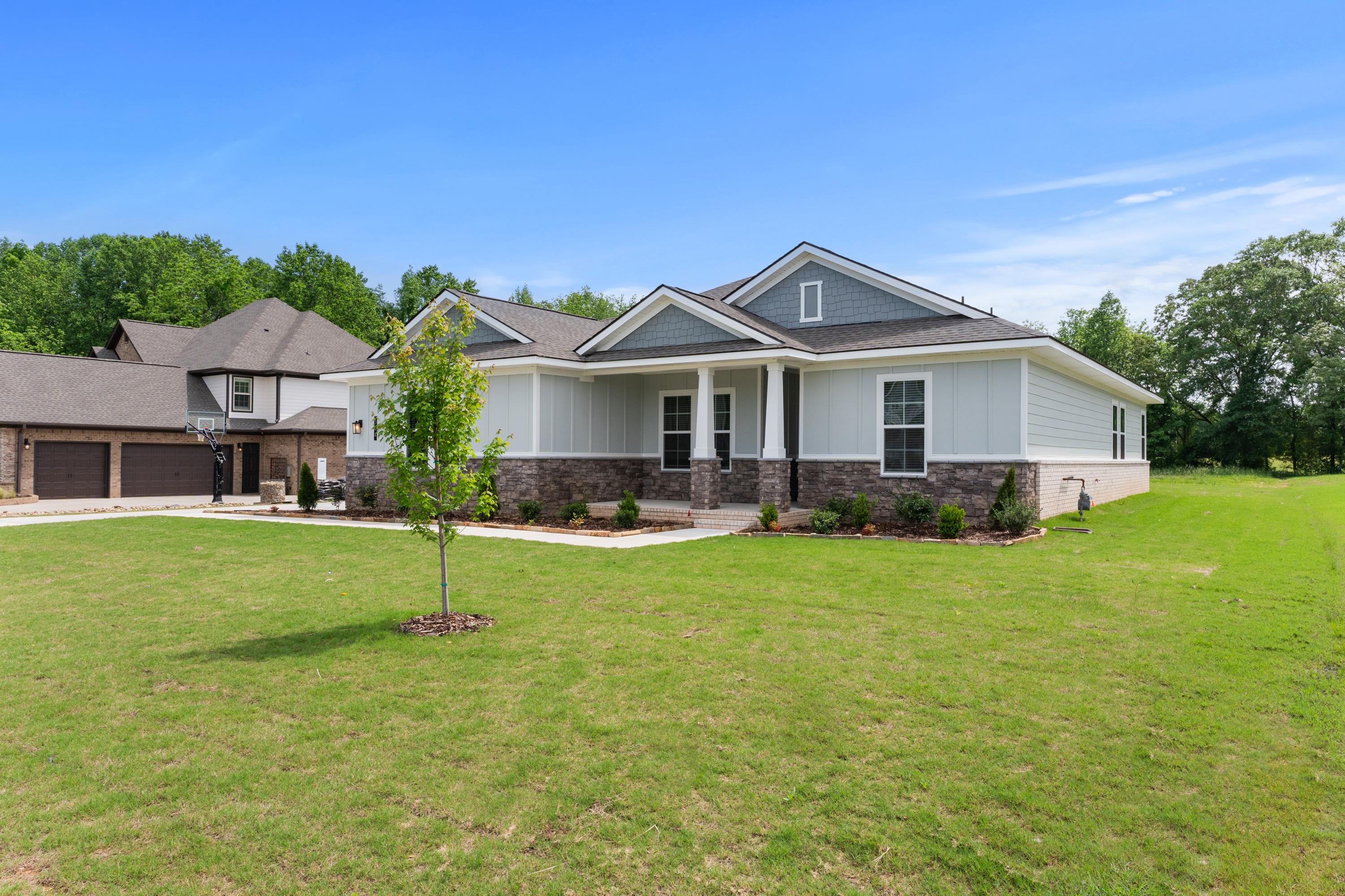 Modern single-story Arcadia B home elevation with gray siding, stone accents, covered porch, 3-car garage, and lush green lawn in Owens Cross Roads