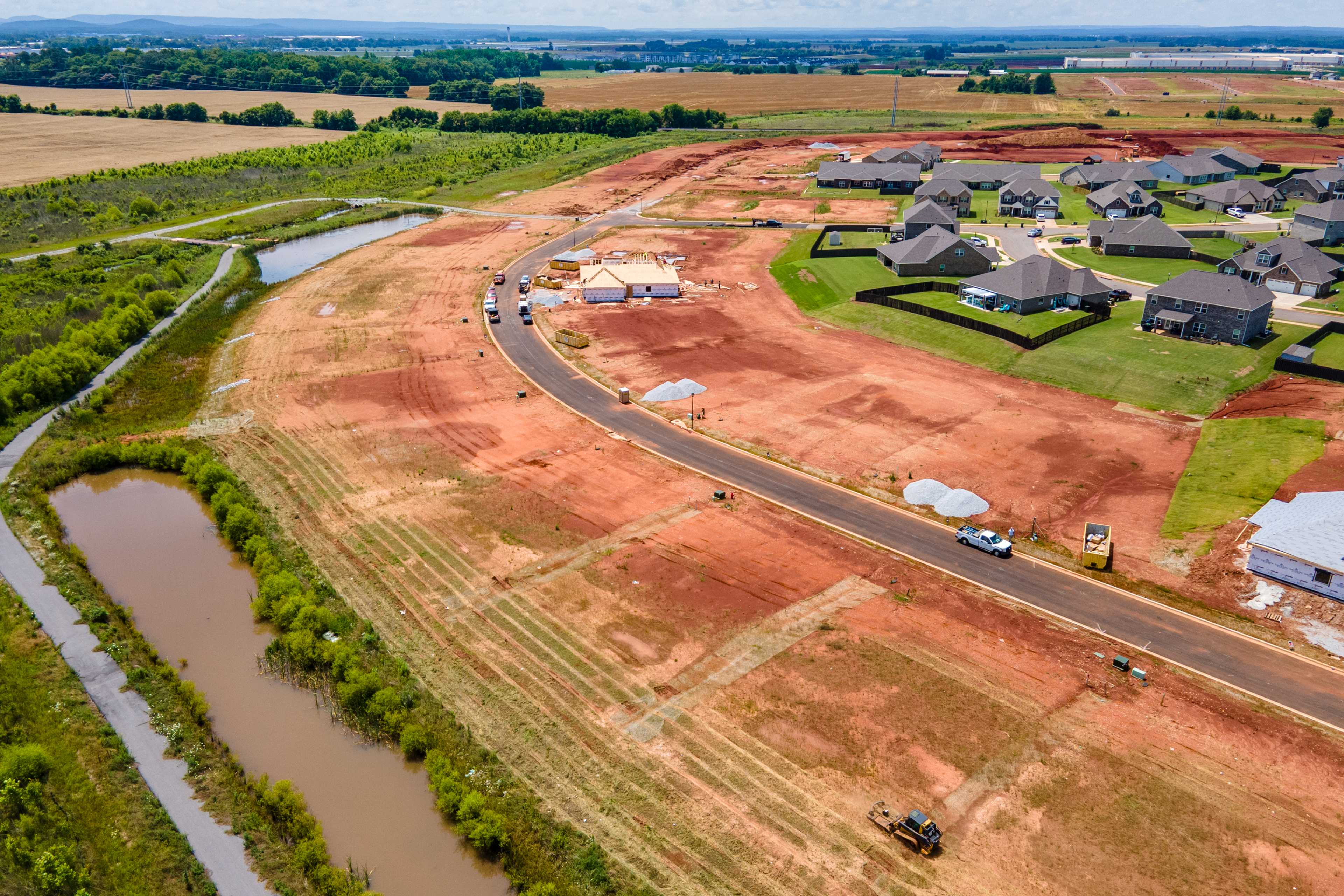 Aerial view of new homes under construction at Barnett's Crossing in Madison, Alabama, with red clay soil, creek, and farmland surroundings