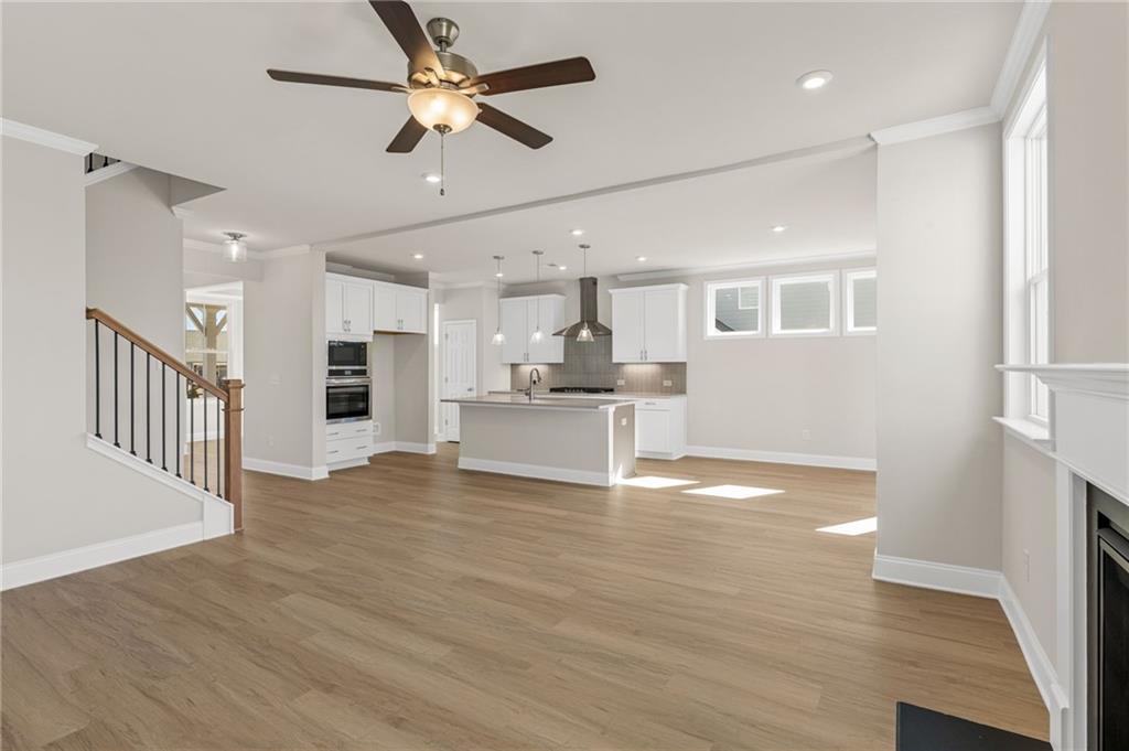 Open-concept living room with white kitchen island, ceiling fan, hardwood floors, and fireplace in The Willow B home, Hoschton, Georgia
