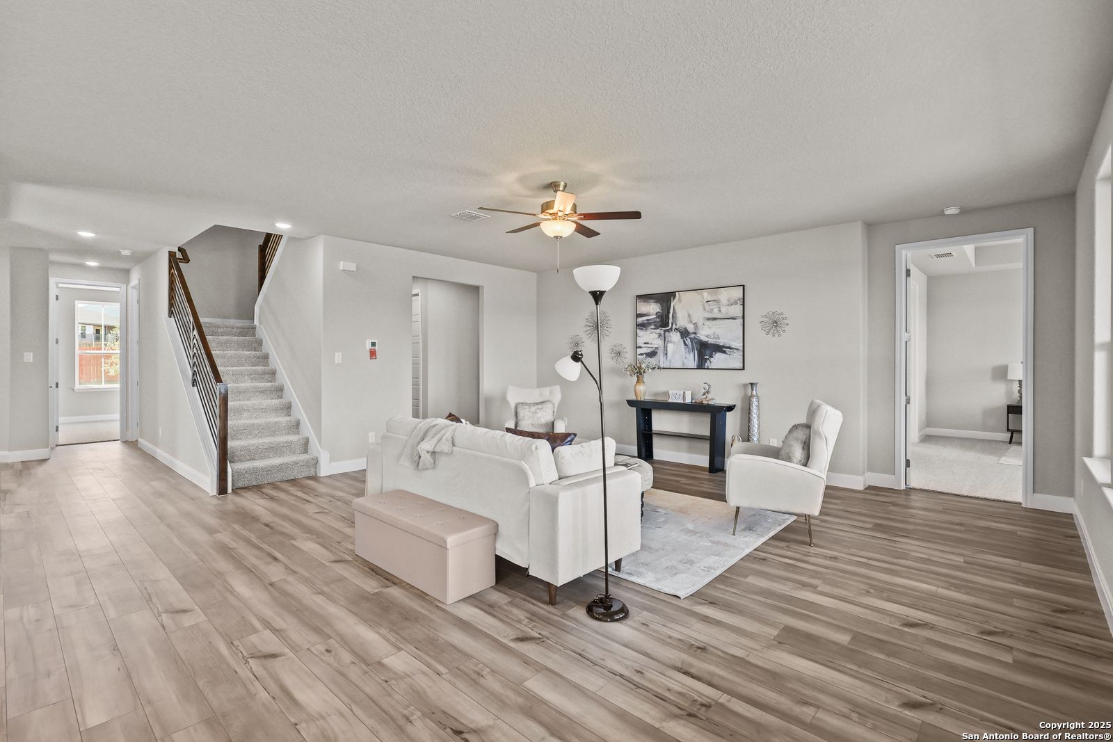 Bright open living room with white sofa, wooden floors, ceiling fan, and staircase in The Jennings G 5-bedroom home, Castroville, Texas