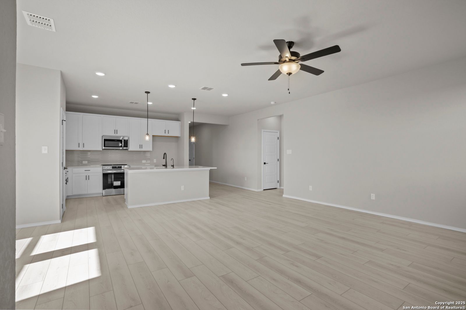 Open-concept kitchen-living area with white cabinets, stainless appliances, island sink, ceiling fan, and hardwood floors in The Daphne H, Seguin, Texas