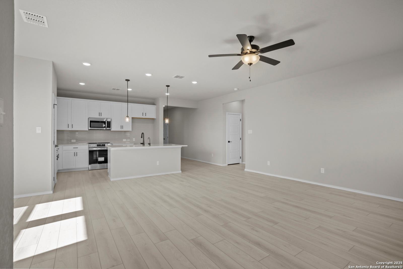 Open-concept kitchen-living area with white cabinets, stainless appliances, island sink, ceiling fan, and hardwood floors in The Daphne H, Seguin, Texas
