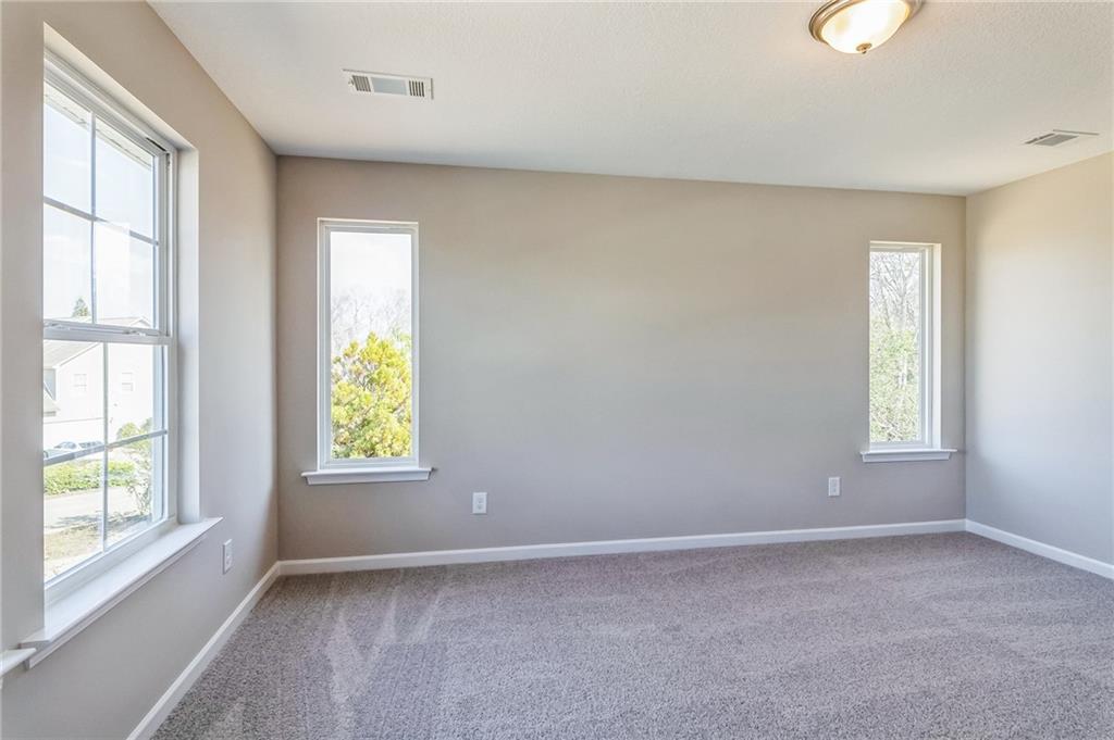 Bright empty bedroom with large windows, beige walls, and carpeted floor in Evermore Homes The Stella, Ivy Glen, Perry, Georgia