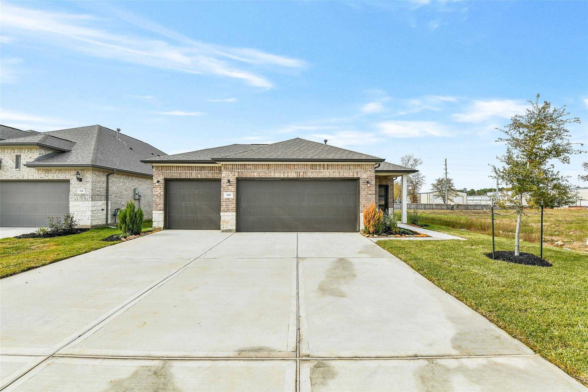 Modern tan brick single-story home with 3-car garage, wide concrete driveway, and landscaped yard in Windmill Estates, Magnolia, Texas