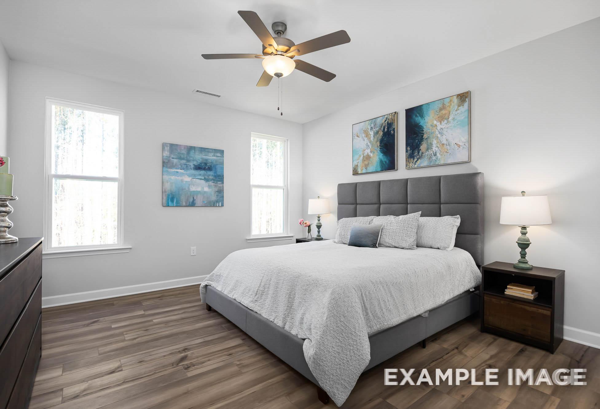 Spacious master bedroom in The Ash with gray upholstered king bed, ceiling fan, abstract wall art, and hardwood floors