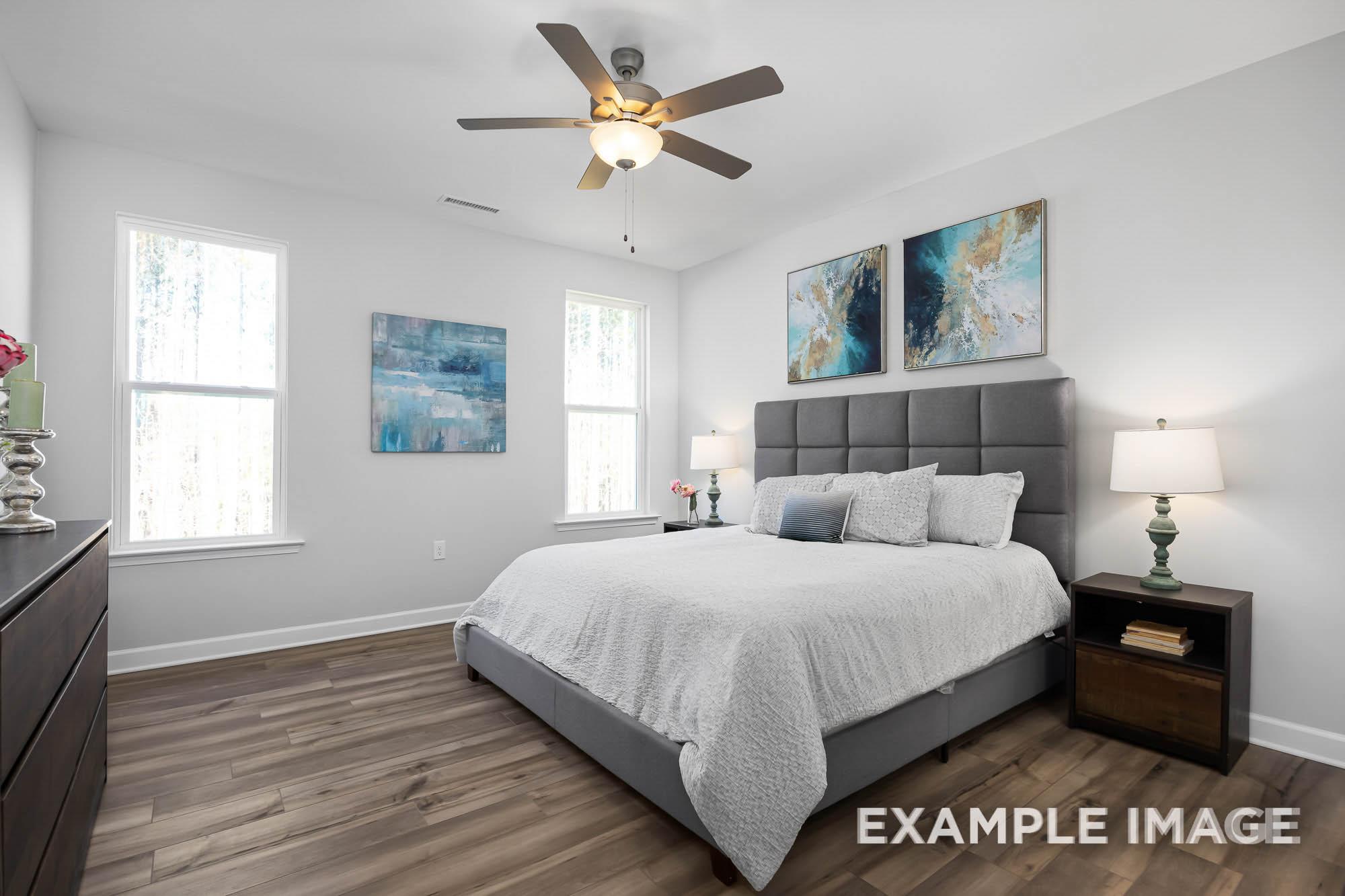 Spacious master bedroom in The Ash with gray upholstered king bed, ceiling fan, abstract wall art, and hardwood floors