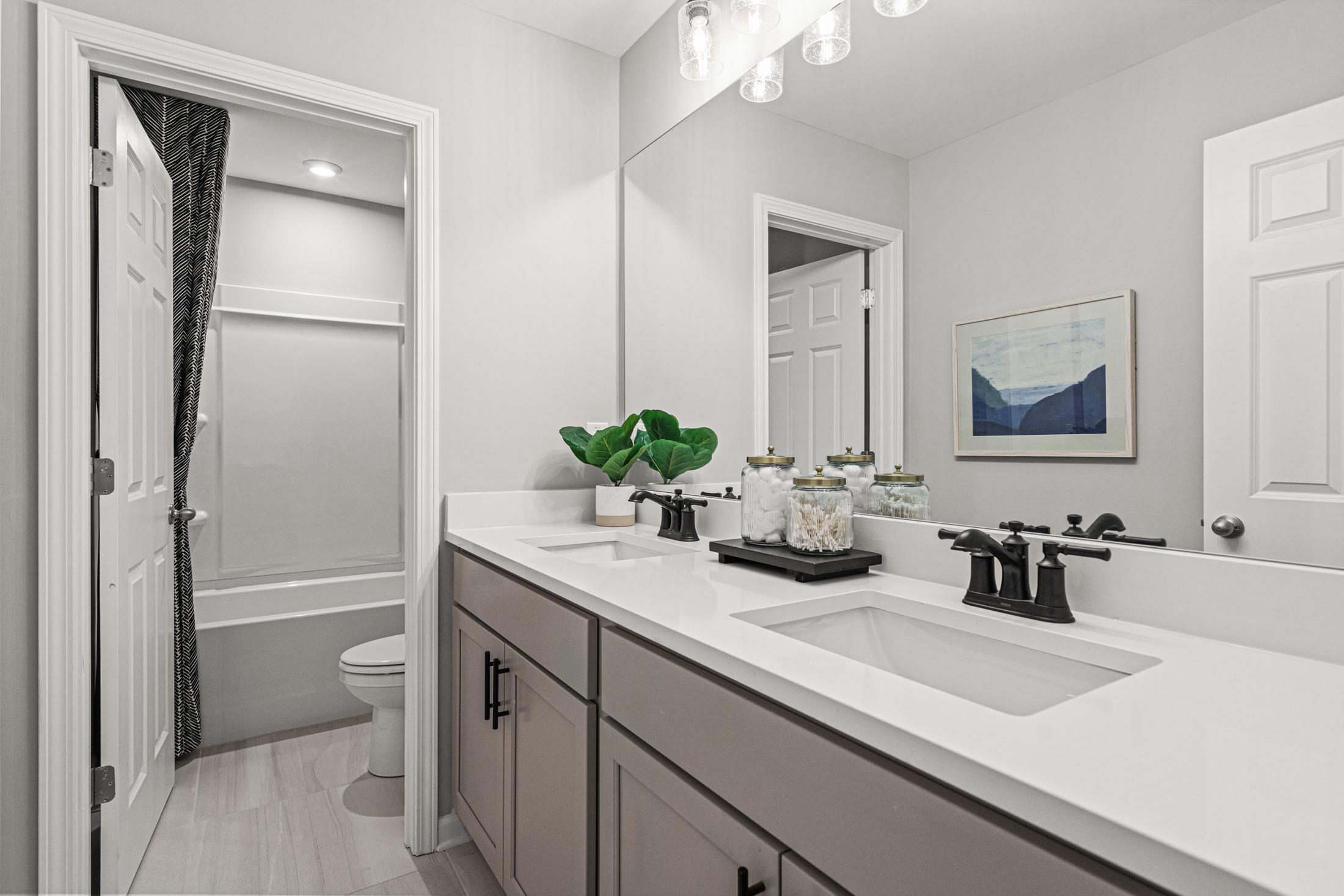 Modern bathroom in The Hickory B showcasing double gray vanity, white quartz countertop, large mirror, potted plants, and adjacent shower tub