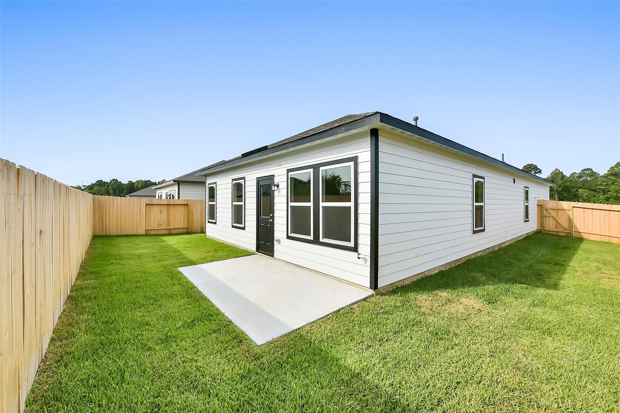 The Comal G single-story 3-bedroom home exterior: white siding, black trim, large windows, green lawn, wooden fence in Dayton, Texas
