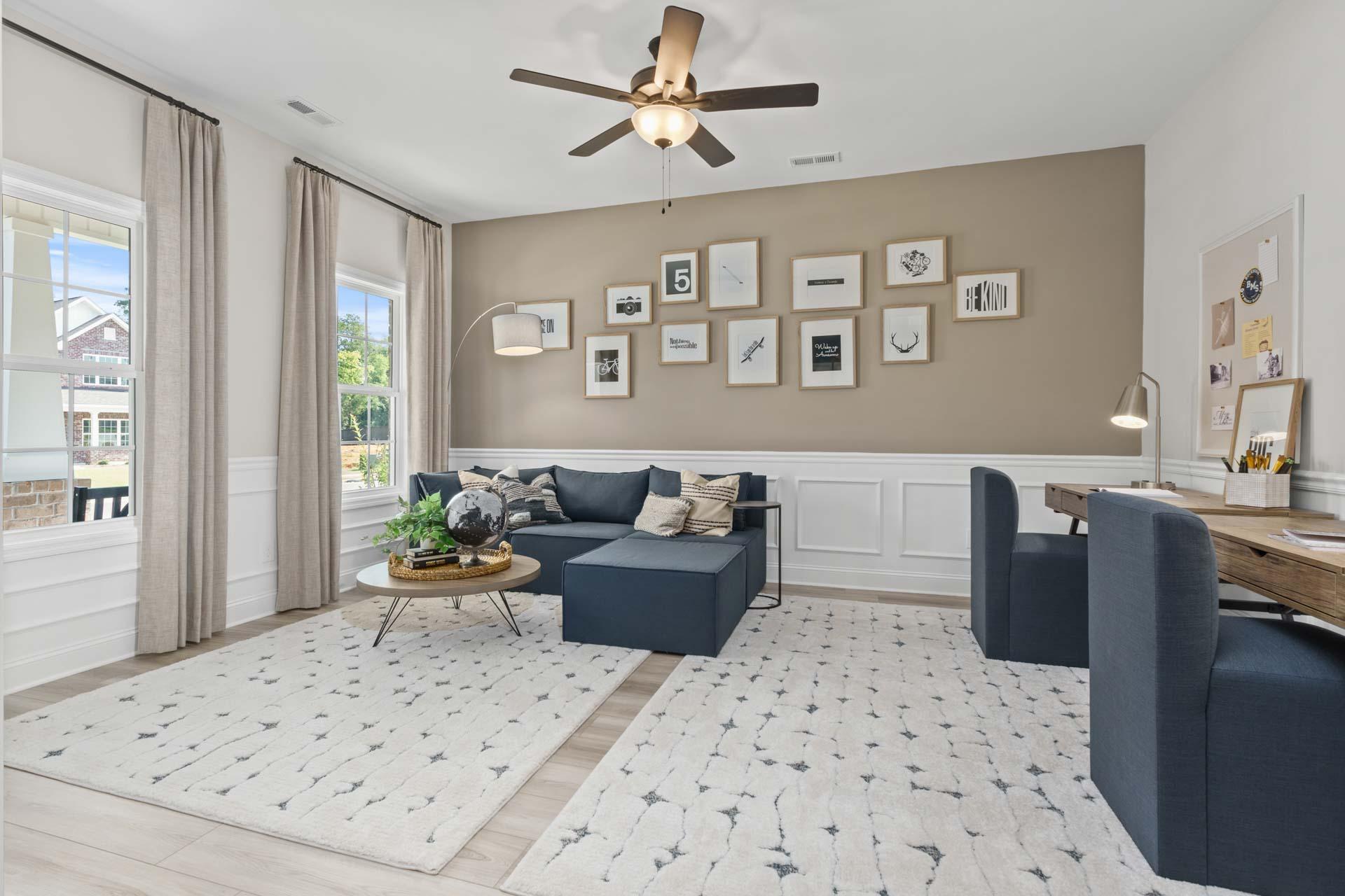 Cozy den in Creek Grove New Market Alabama by Davidson Homes featuring blue sectional sofa, desk, wall art gallery, and large windows