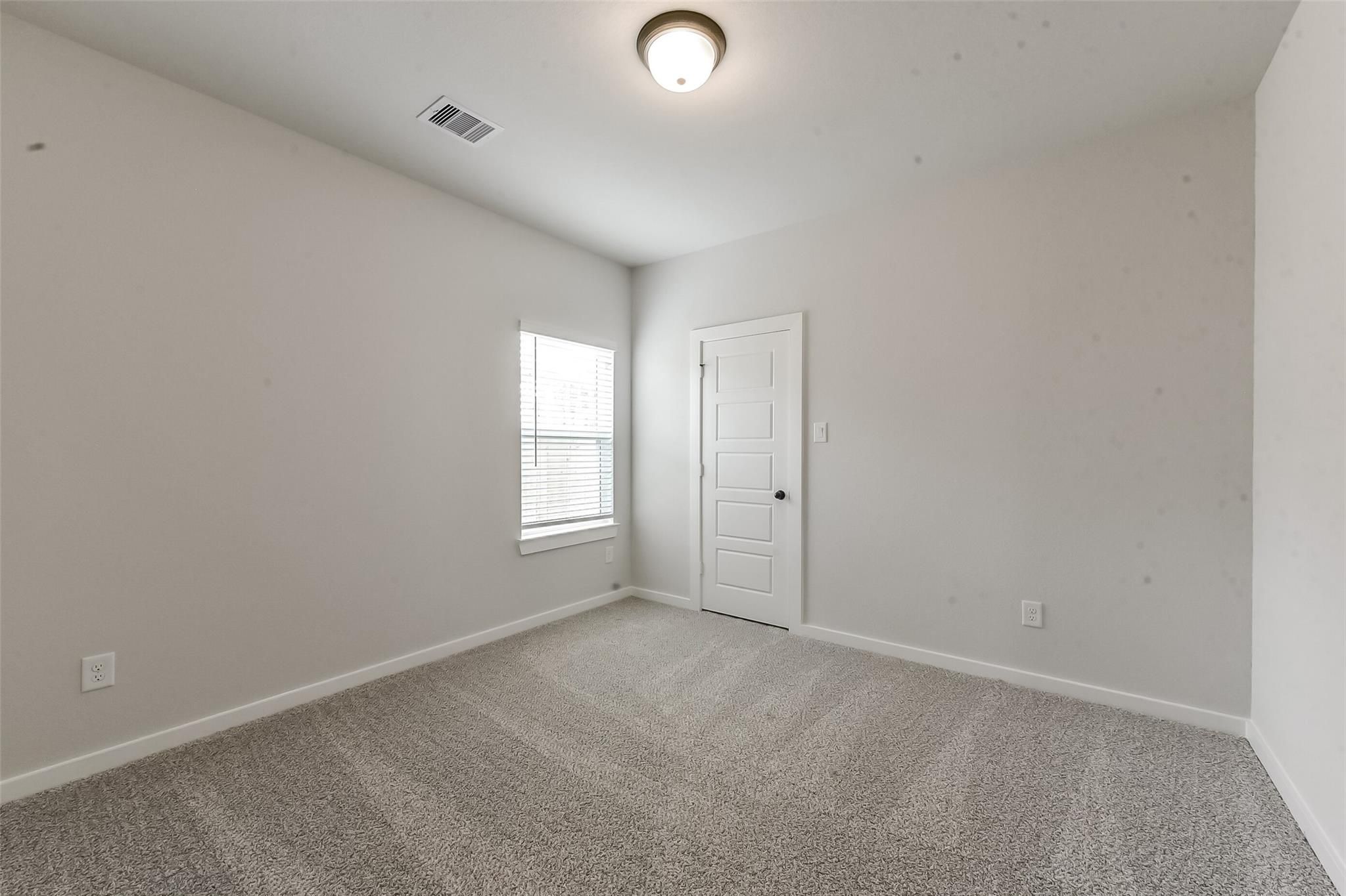 Empty secondary bedroom featuring light gray walls, plush gray carpet, window with blinds, and white door in Davidson Homes The Everett C, Crosby Texas