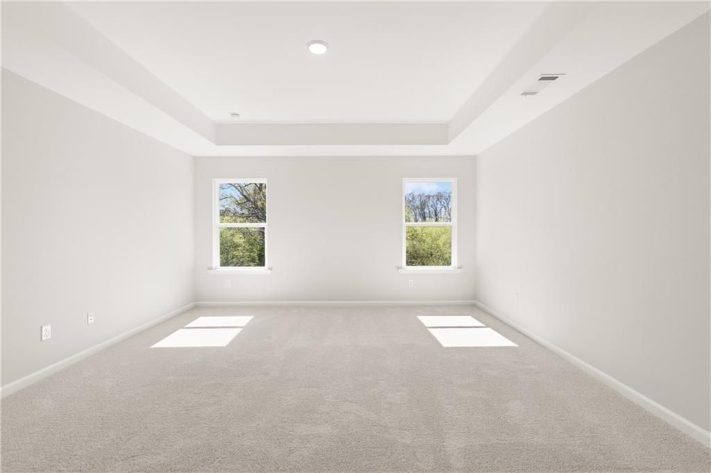 Bright empty bedroom with tray ceiling, light gray carpet, and large windows to greenery in The Hickory B, Winder, Georgia