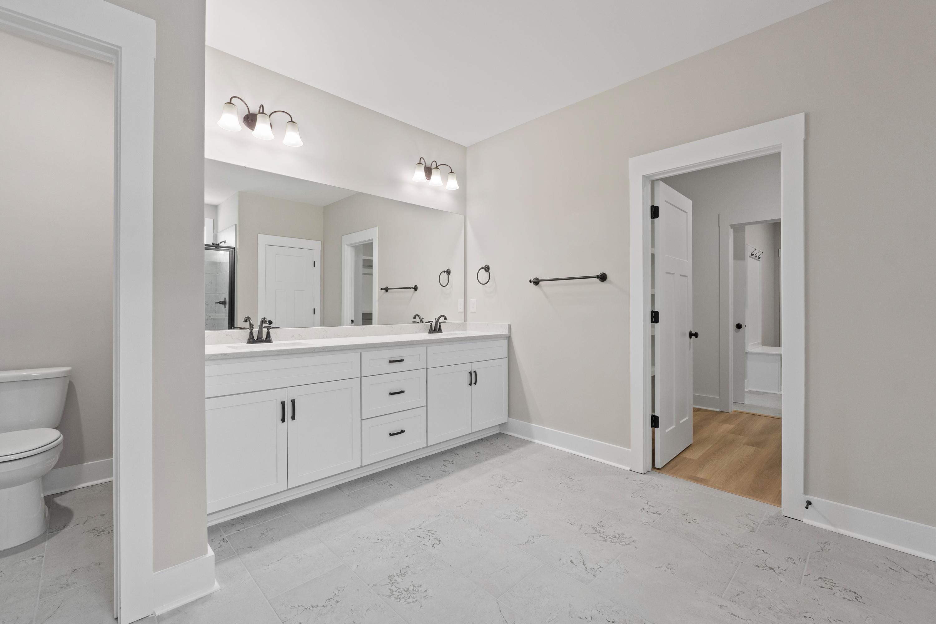 Spacious master bathroom in The Oxford with double white vanity, large mirrors, sconce lights, toilet, and adjacent doorway