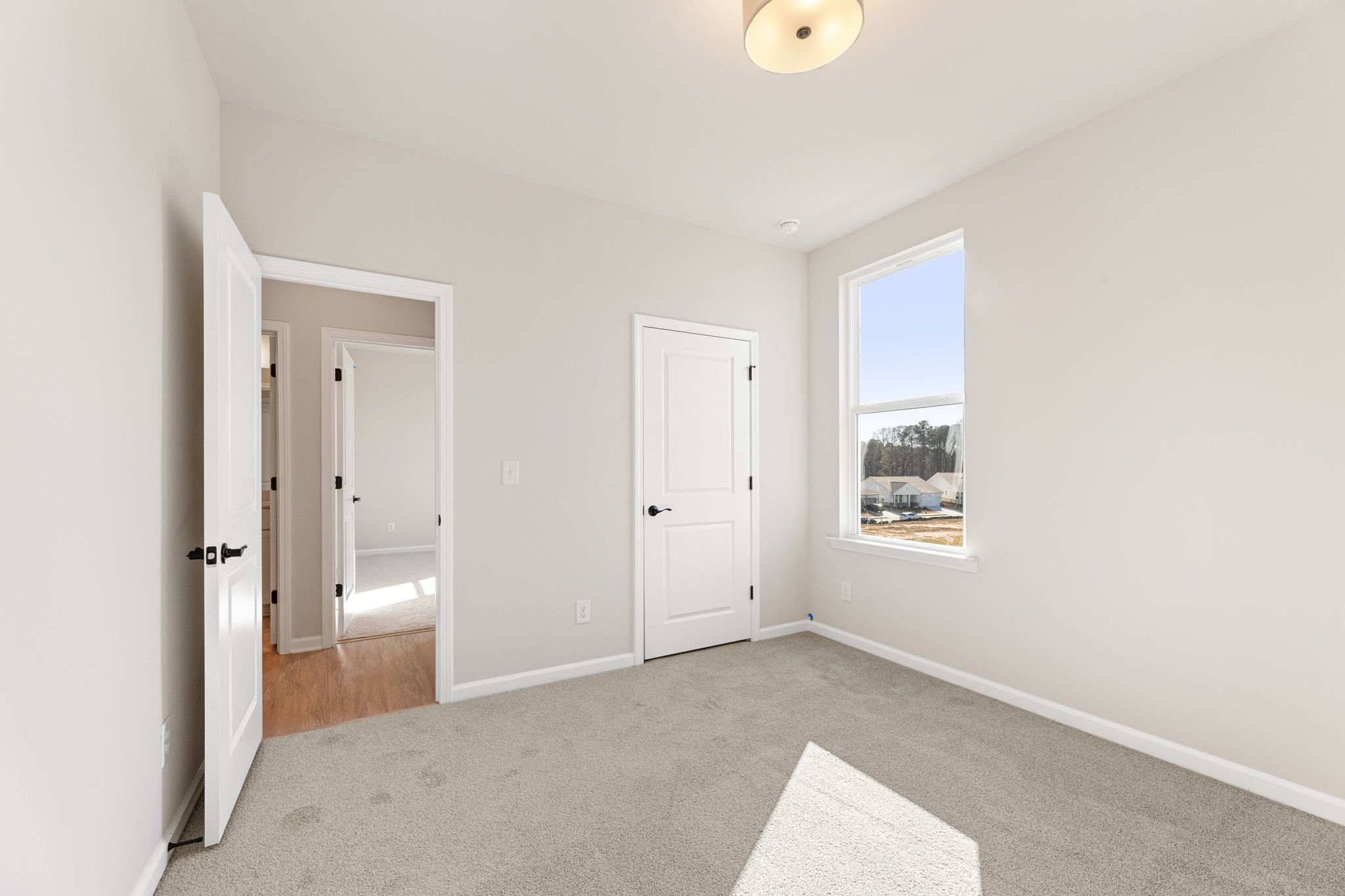 Bright secondary bedroom with beige walls, gray carpet, adjacent bath door, and sunny window in Davidson Homes The Glenwood C, Loganville, GA