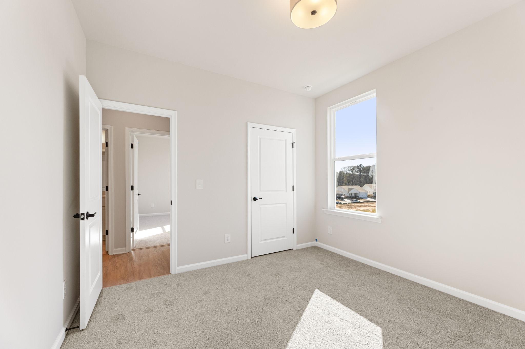 Bright secondary bedroom with beige walls, gray carpet, adjacent bath door, and sunny window in Davidson Homes The Glenwood C, Loganville, GA