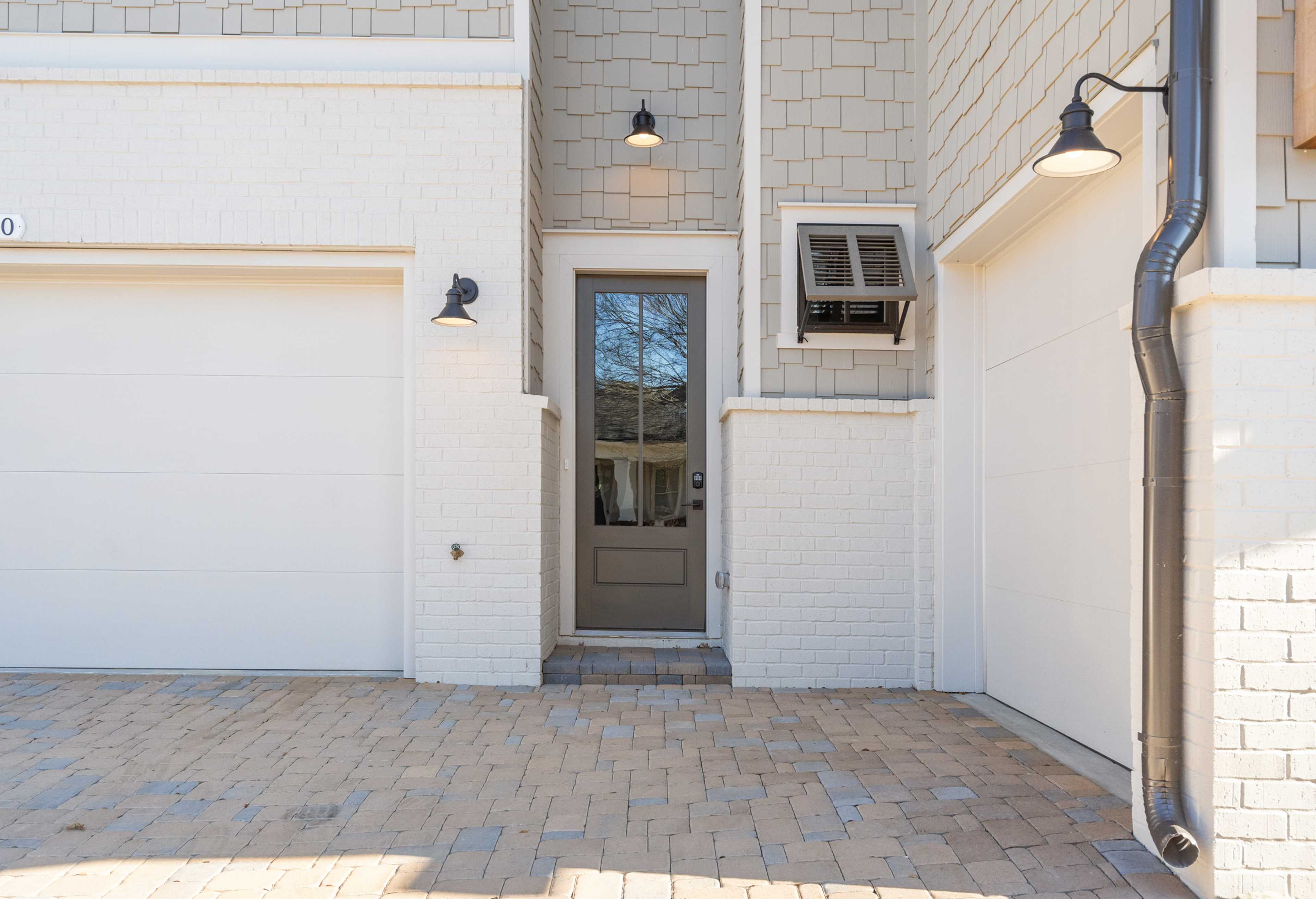 The Seaside 3-car garage facade with white siding, gray entry door, wall lanterns, shutters, and paver driveway in Woodstock, Georgia