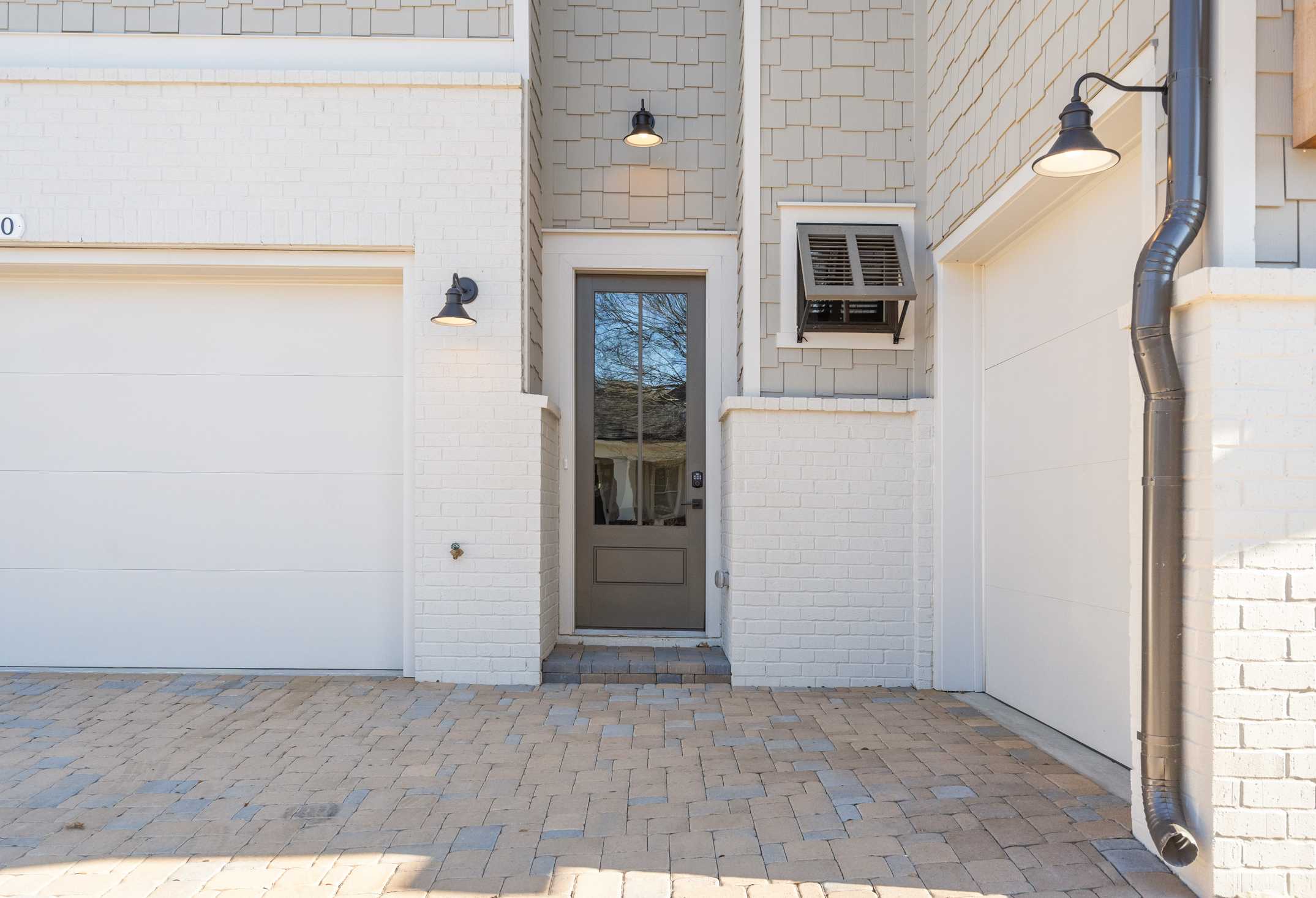 The Seaside 3-car garage facade with white siding, gray entry door, wall lanterns, shutters, and paver driveway in Woodstock, Georgia