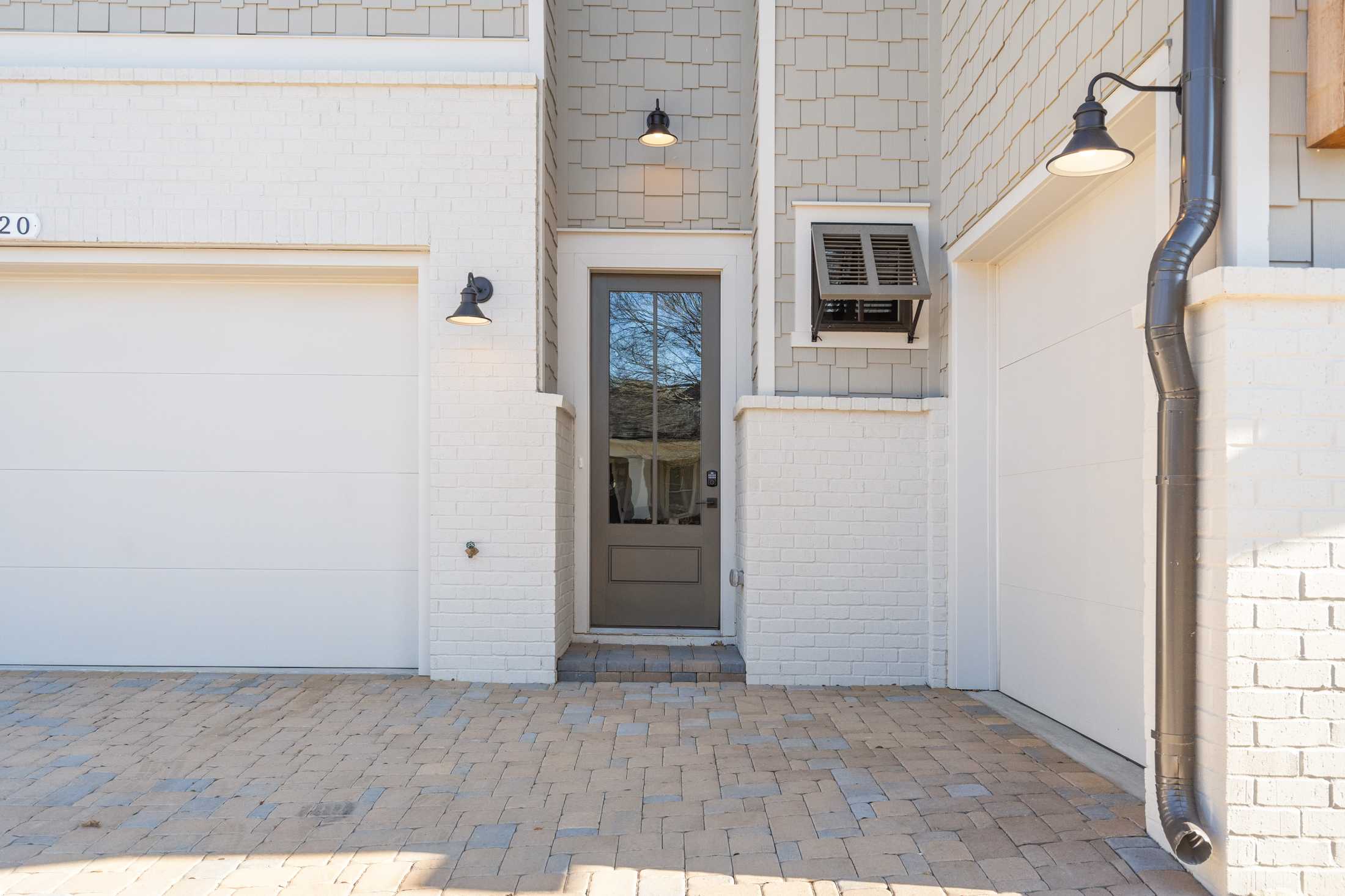 The Seaside 3-car garage facade with white siding, gray entry door, wall lanterns, shutters, and paver driveway in Woodstock, Georgia