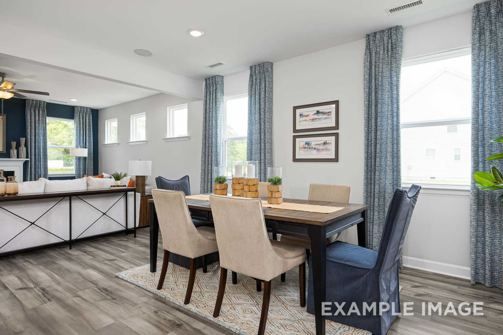 Spacious dining room in The Adalynn A with wooden table, beige upholstered chairs, blue curtains, and open living area