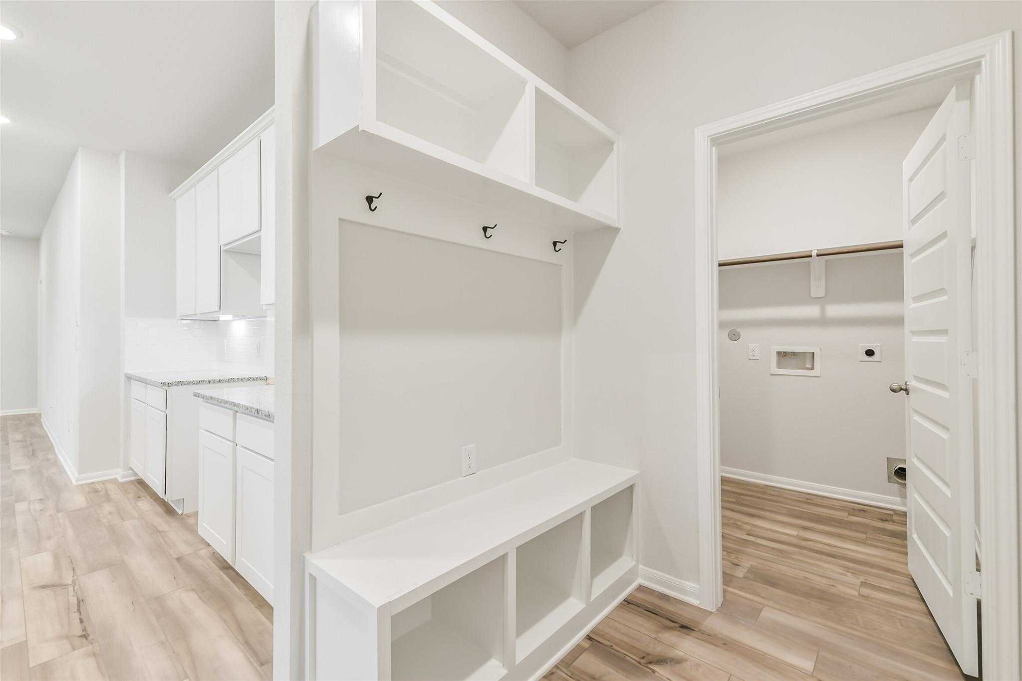 White mudroom with built-in bench, coat hooks, open shelves, and adjacent laundry room in Davidson Homes The Brazos F, Conroe, Texas