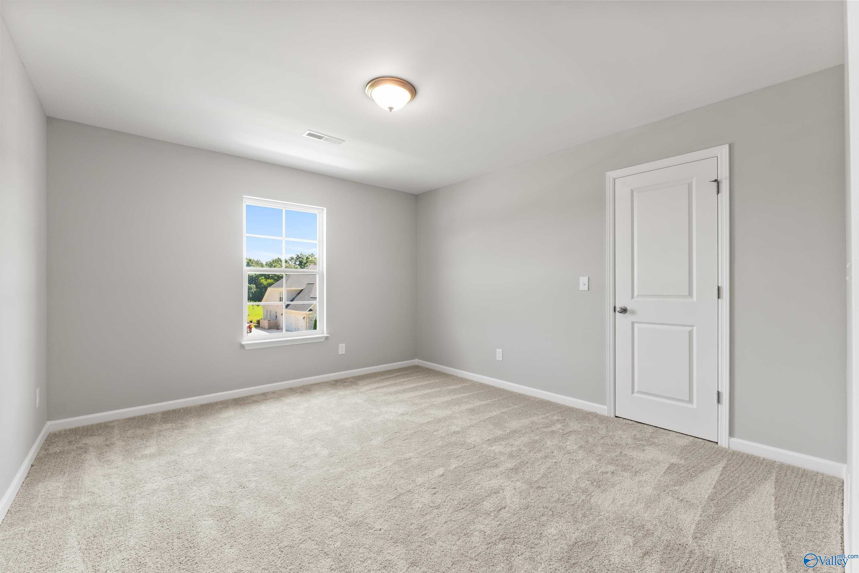 Bright secondary bedroom with light gray walls, neutral carpet, white door and window overlooking trees in The Charm home, Huntsville