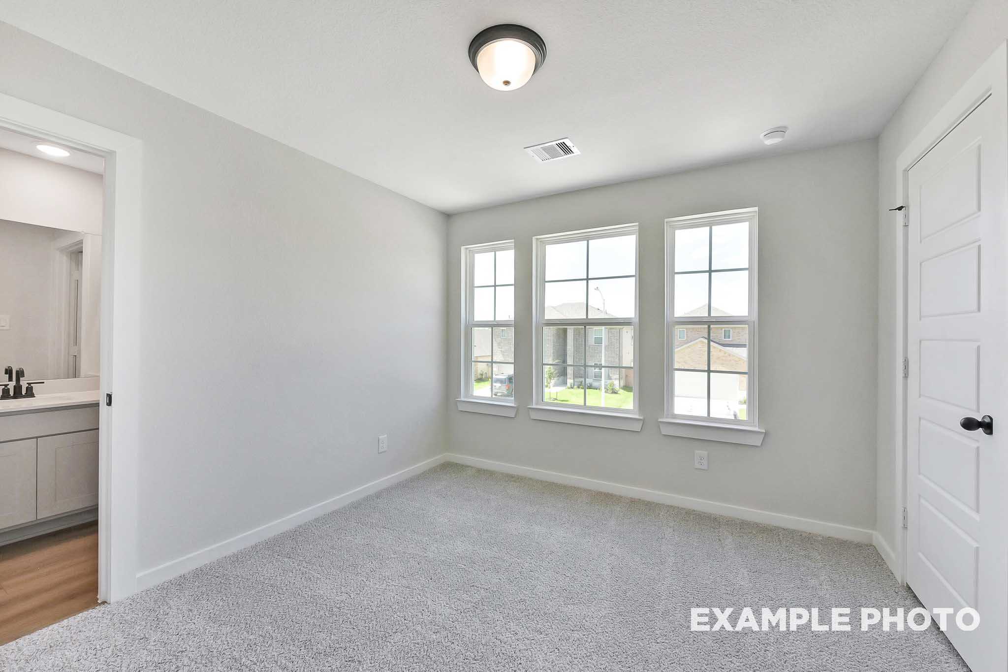 Bright secondary bedroom in The Sequoia Davidson Homes plan, gray walls, carpet flooring, three large windows, en-suite bathroom with vanity