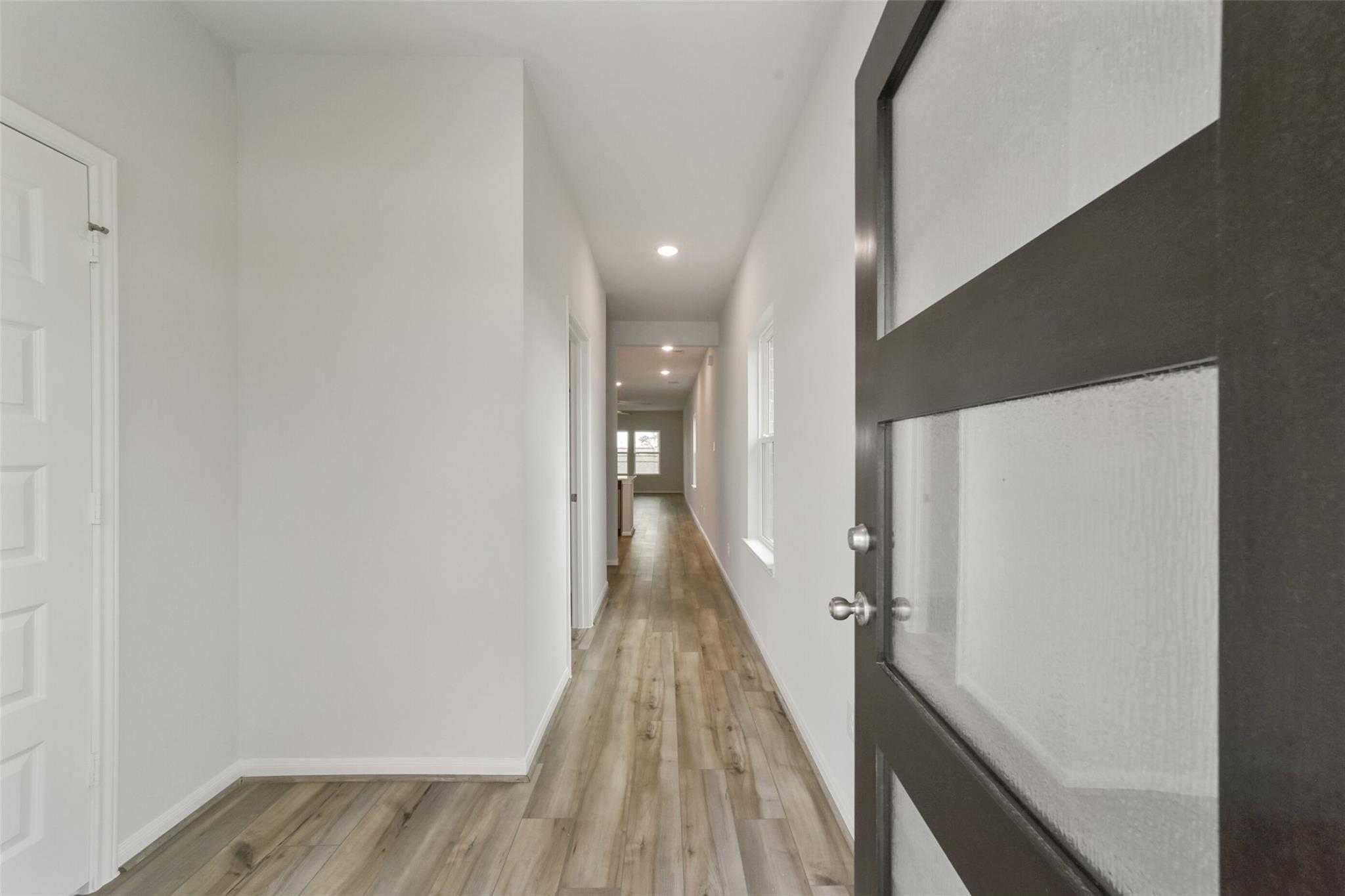 Long hallway with light hardwood floors, white walls, and frosted glass entry door in Davidson Homes San Marcos E, Beasley, Texas