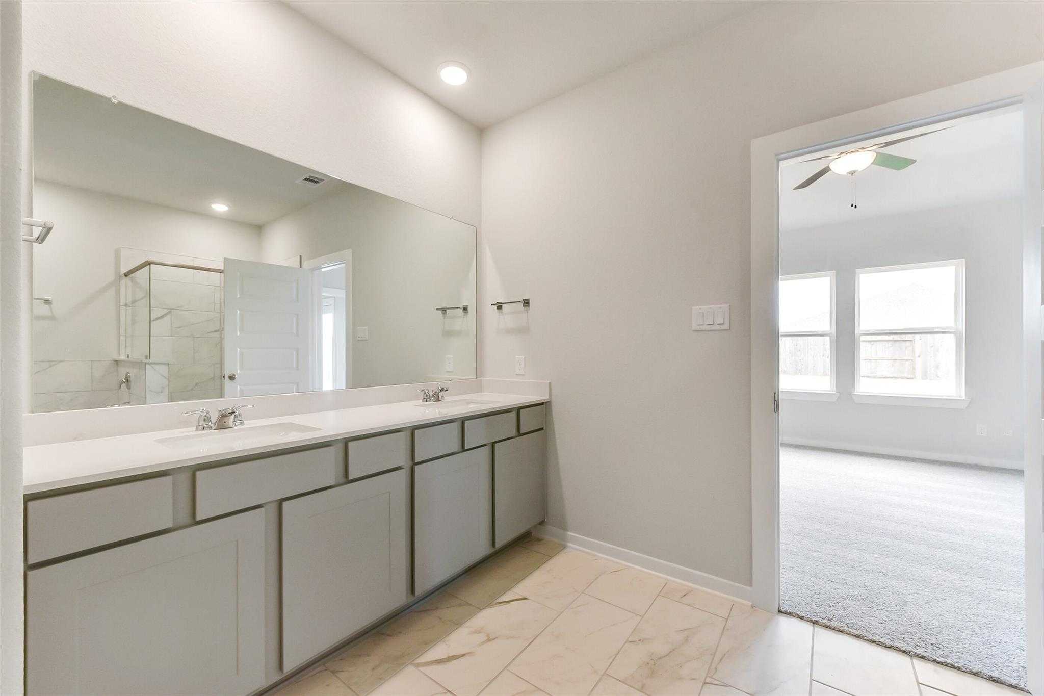 Modern master bathroom with double vanity, frameless glass shower, and adjacent bedroom in Davidson Homes The Laguna C, Rosharon, Texas