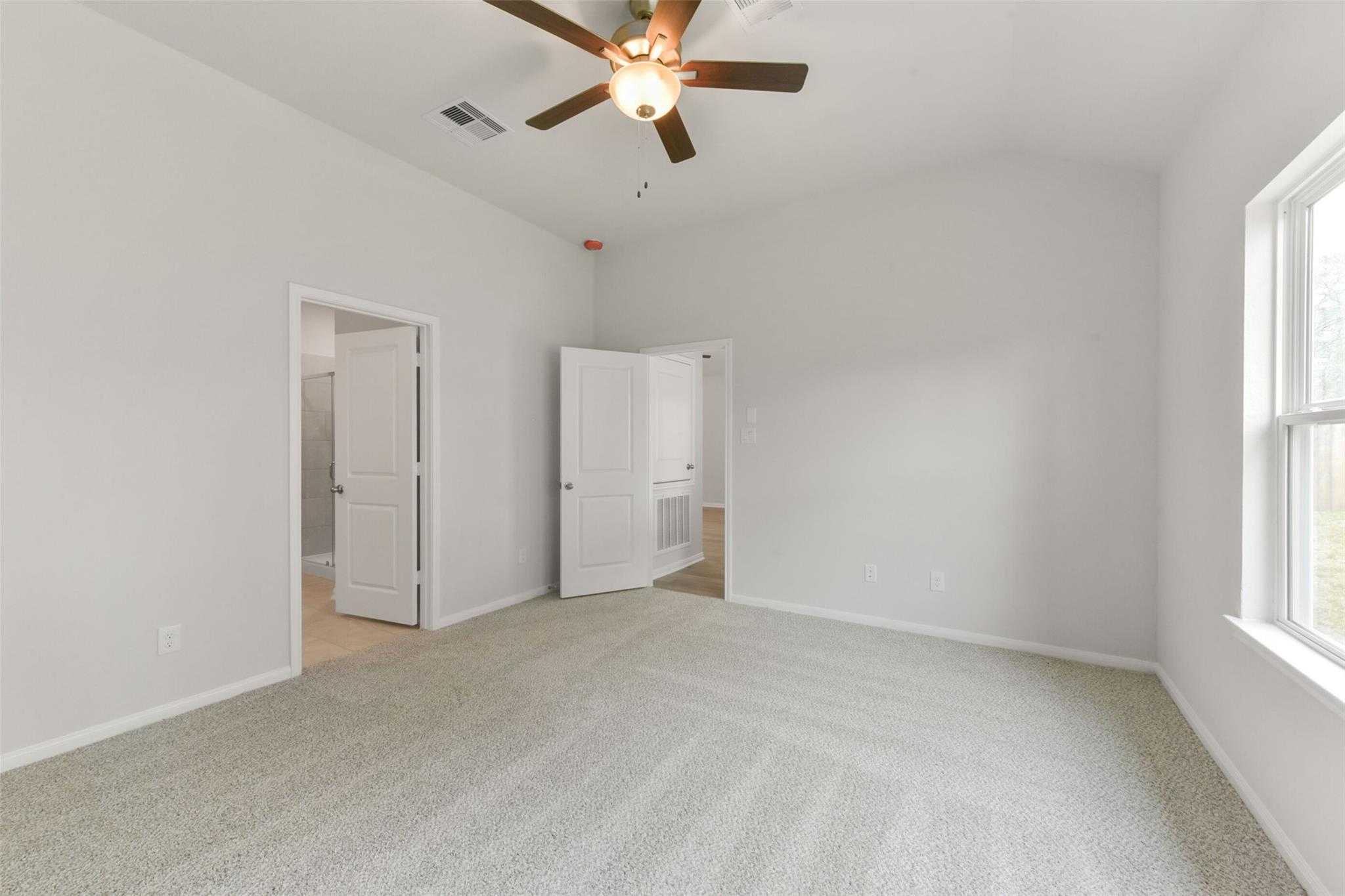Bright bedroom with ceiling fan, beige carpet, open bathroom door, and window in The Laguna B, Magnolia, Texas