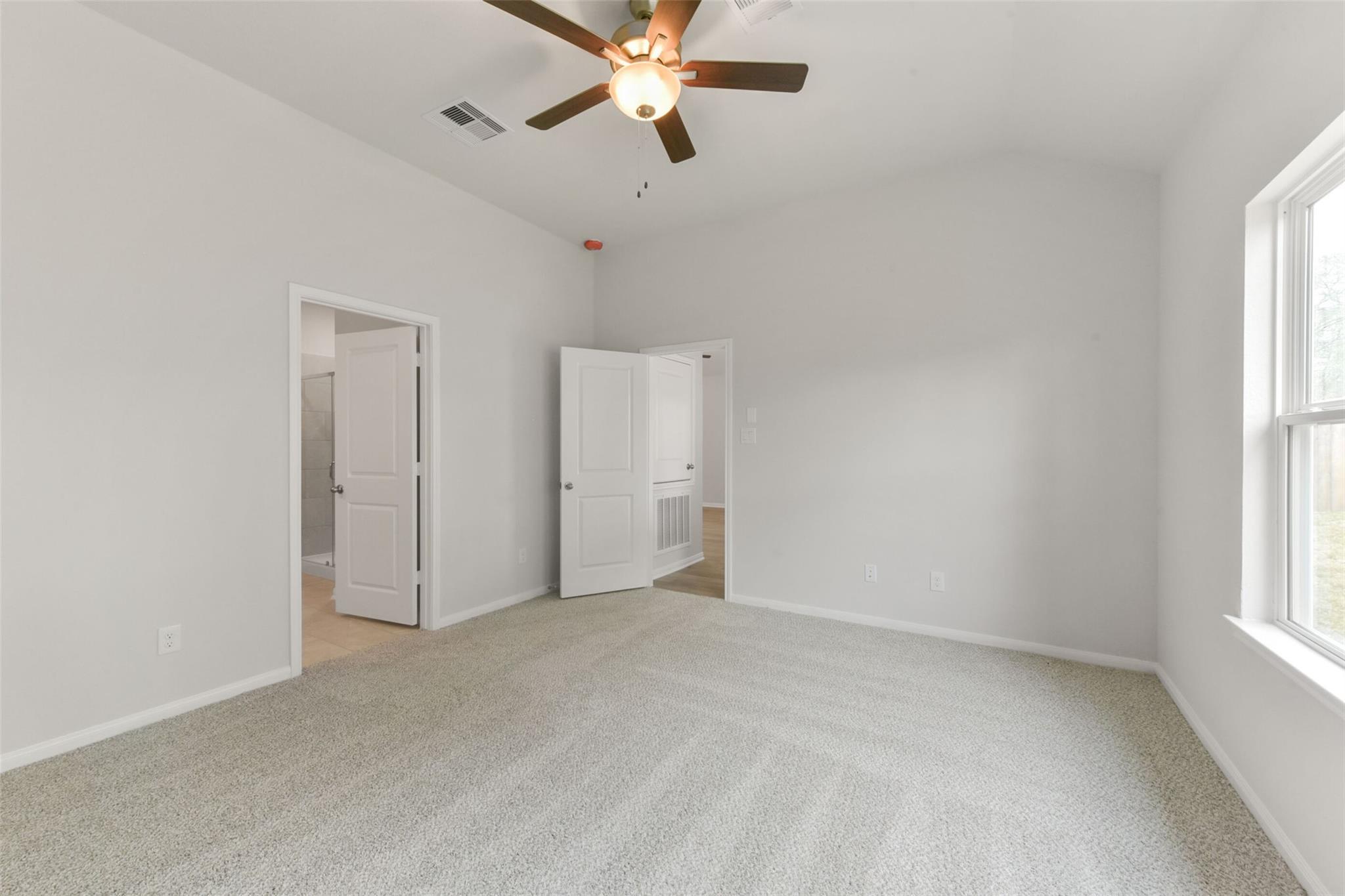 Bright bedroom with ceiling fan, beige carpet, open bathroom door, and window in The Laguna B, Magnolia, Texas
