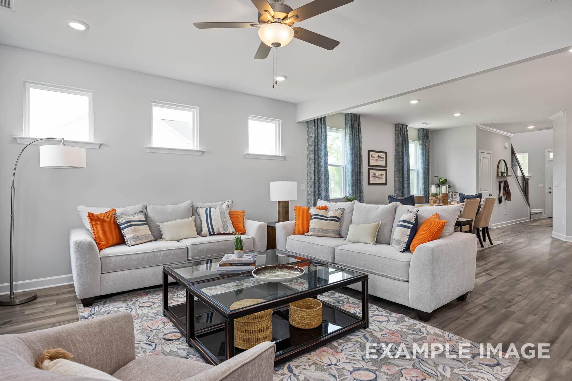Cozy living room in The Adalynn C Davidson Homes with neutral gray sofas, orange pillows, glass coffee table, ceiling fan, and open dining area