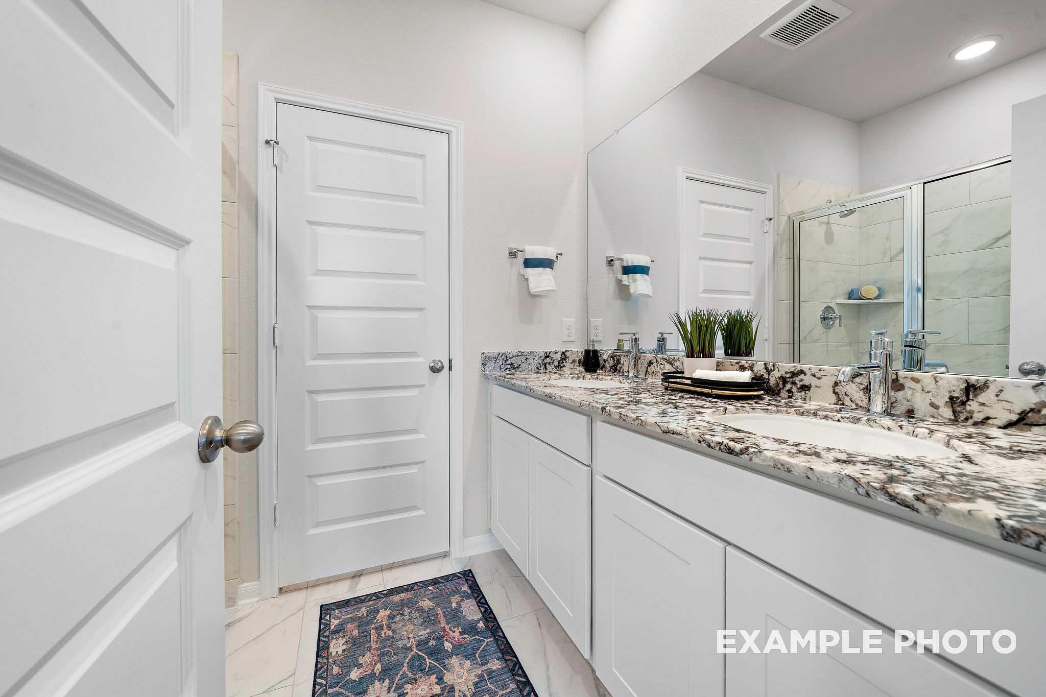Spacious master bathroom in The San Marcos plan featuring white cabinetry, granite vanity, dual sinks, and frameless glass shower
