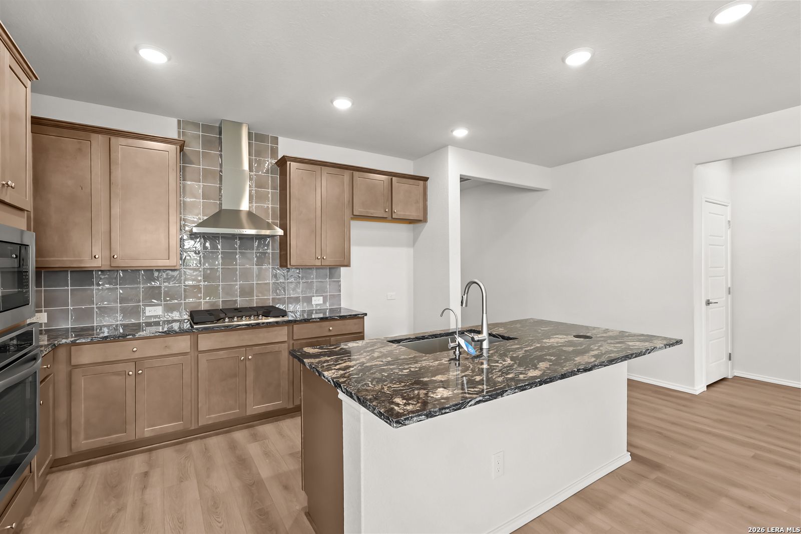 Kitchen with granite island, stainless gas cooktop, tile backsplash, and wood-look flooring in Davidson Homes The Daphne I, San Antonio