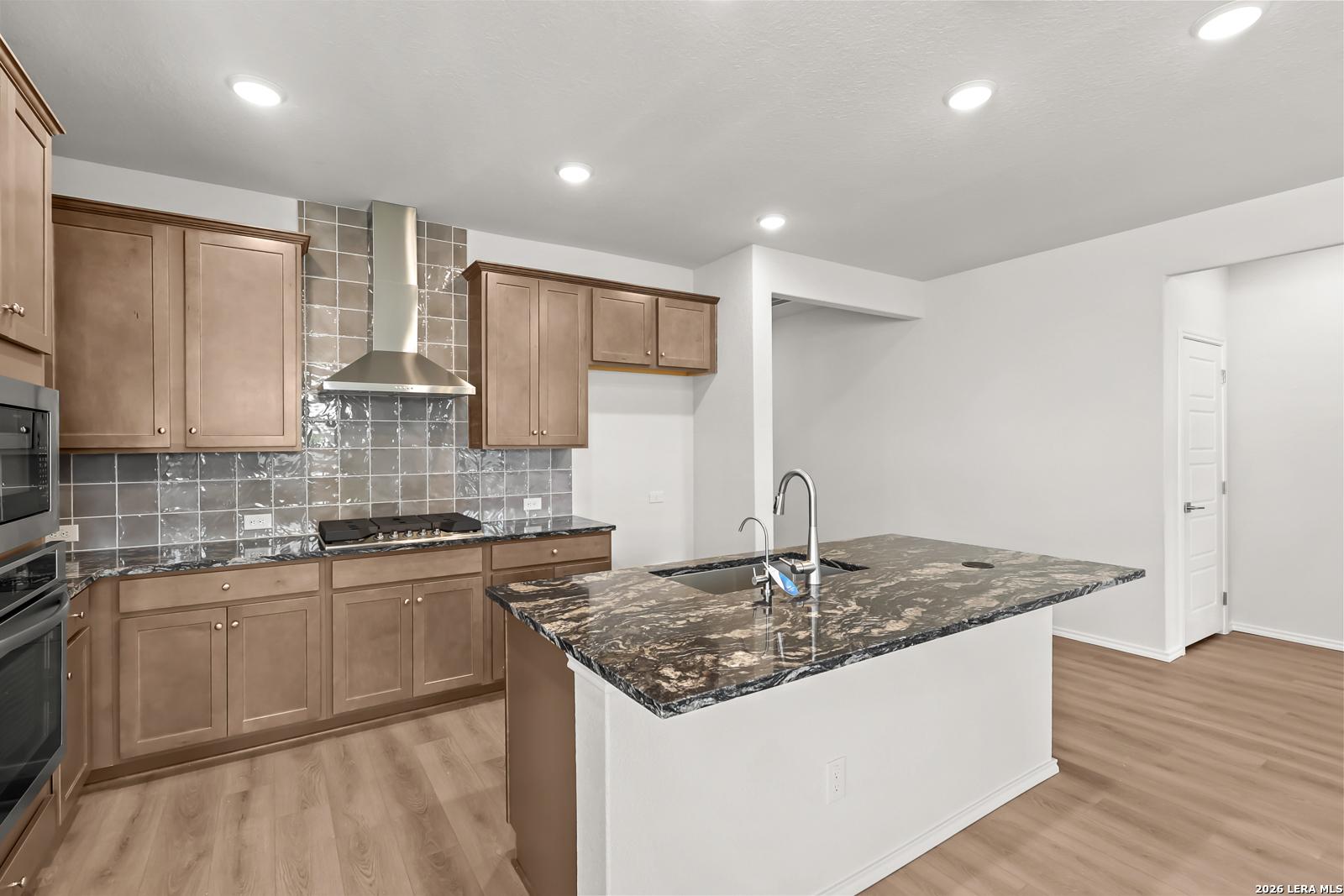 Kitchen with granite island, stainless gas cooktop, tile backsplash, and wood-look flooring in Davidson Homes The Daphne I, San Antonio