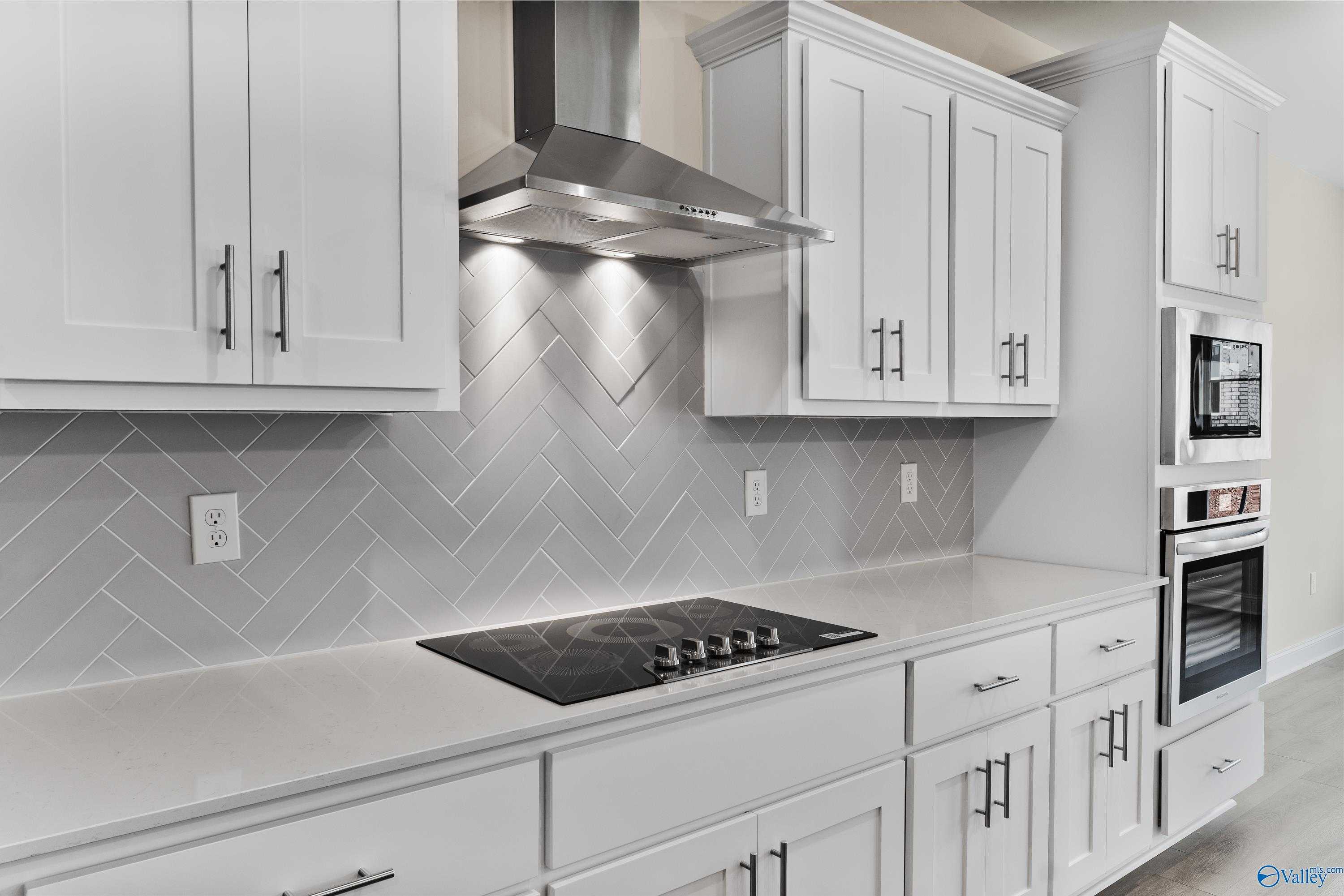 Modern white kitchen featuring gray herringbone backsplash, stainless range hood, induction cooktop in The Finleigh by Davidson Homes, Toney, AL