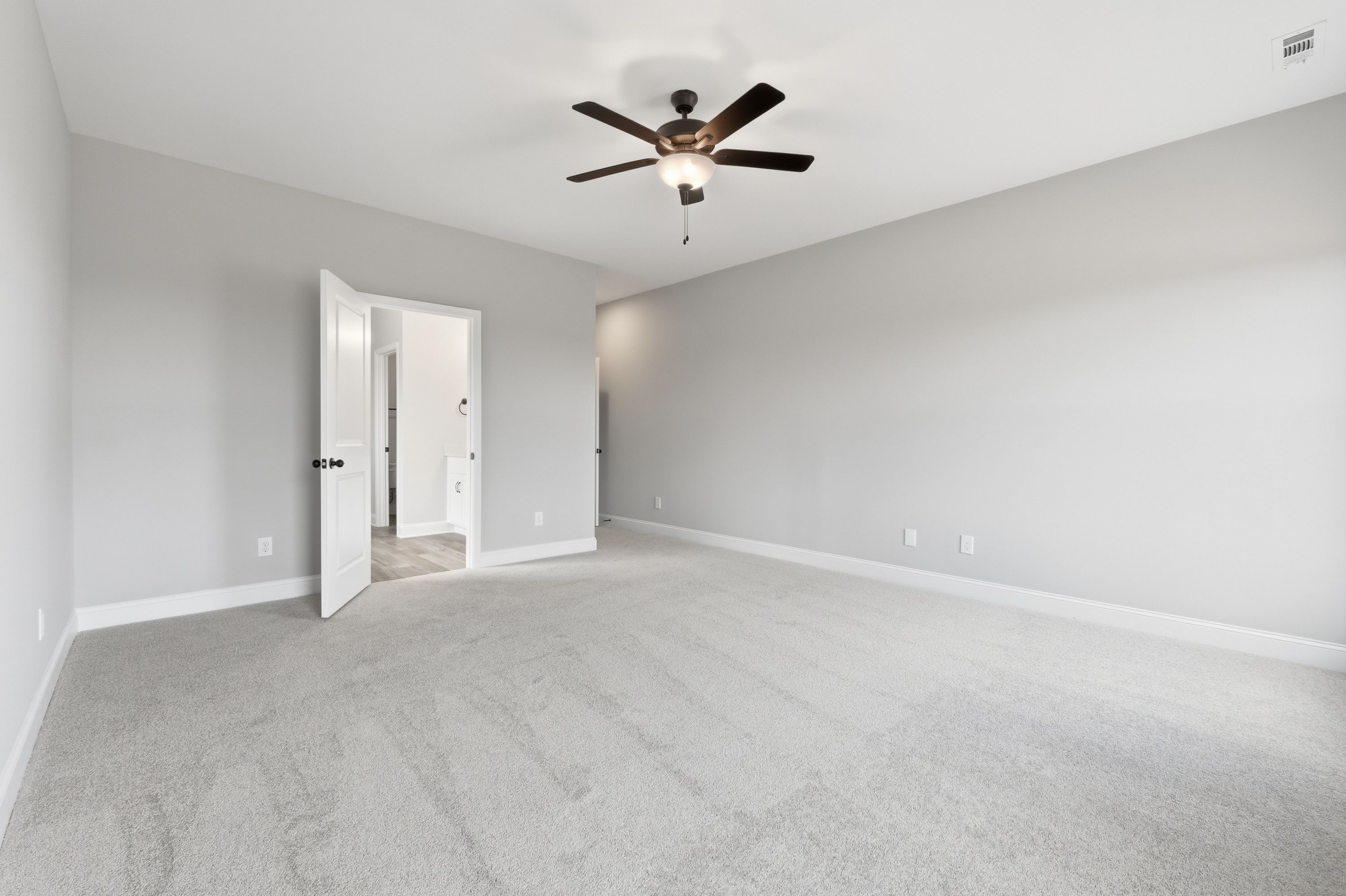 Spacious master bedroom in The Rockford home with gray carpet, ceiling fan, and open door to en-suite bath