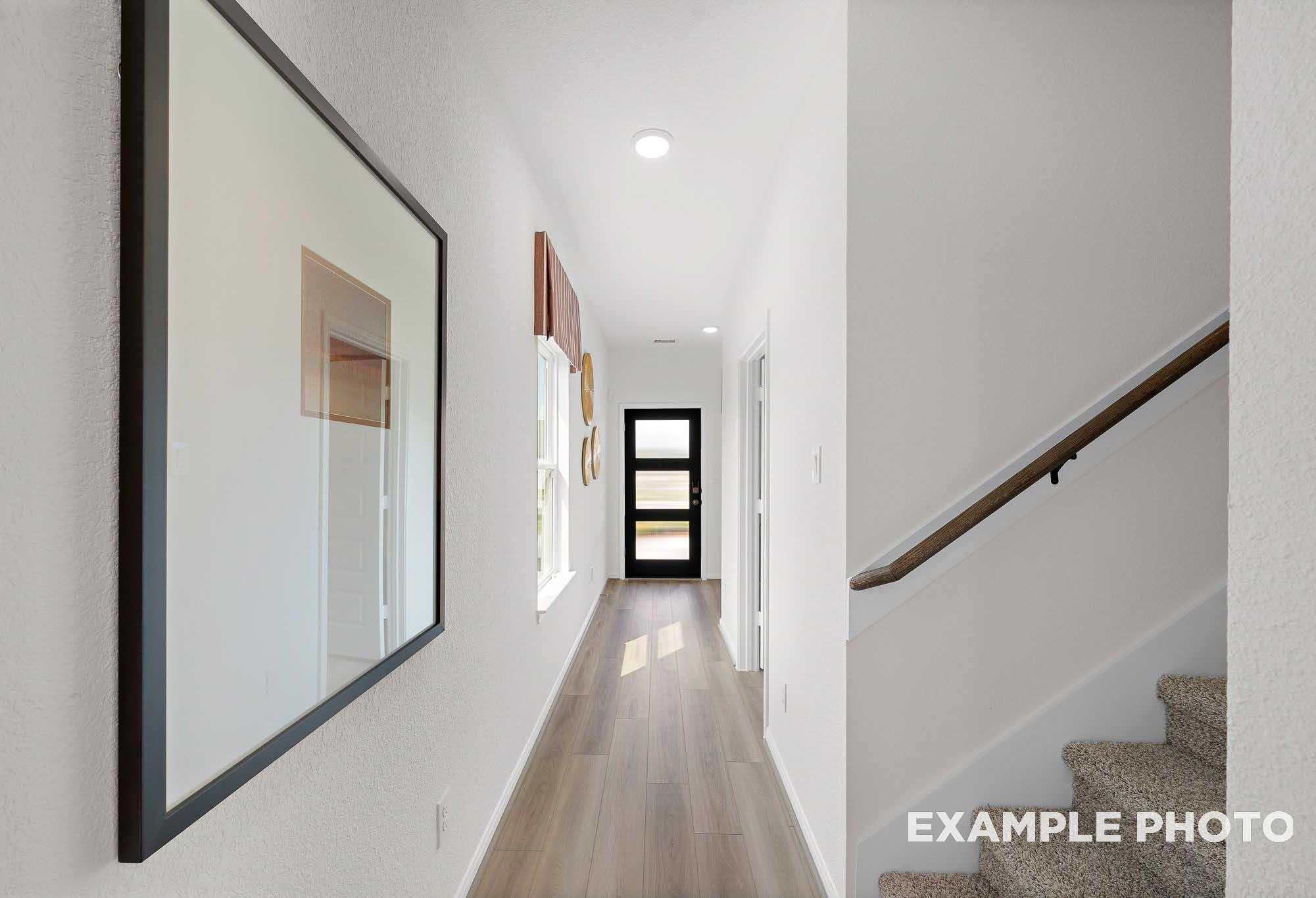 Spacious hallway in The Sabine home with oak hardwood floors, white walls, modern black door, and carpeted staircase
