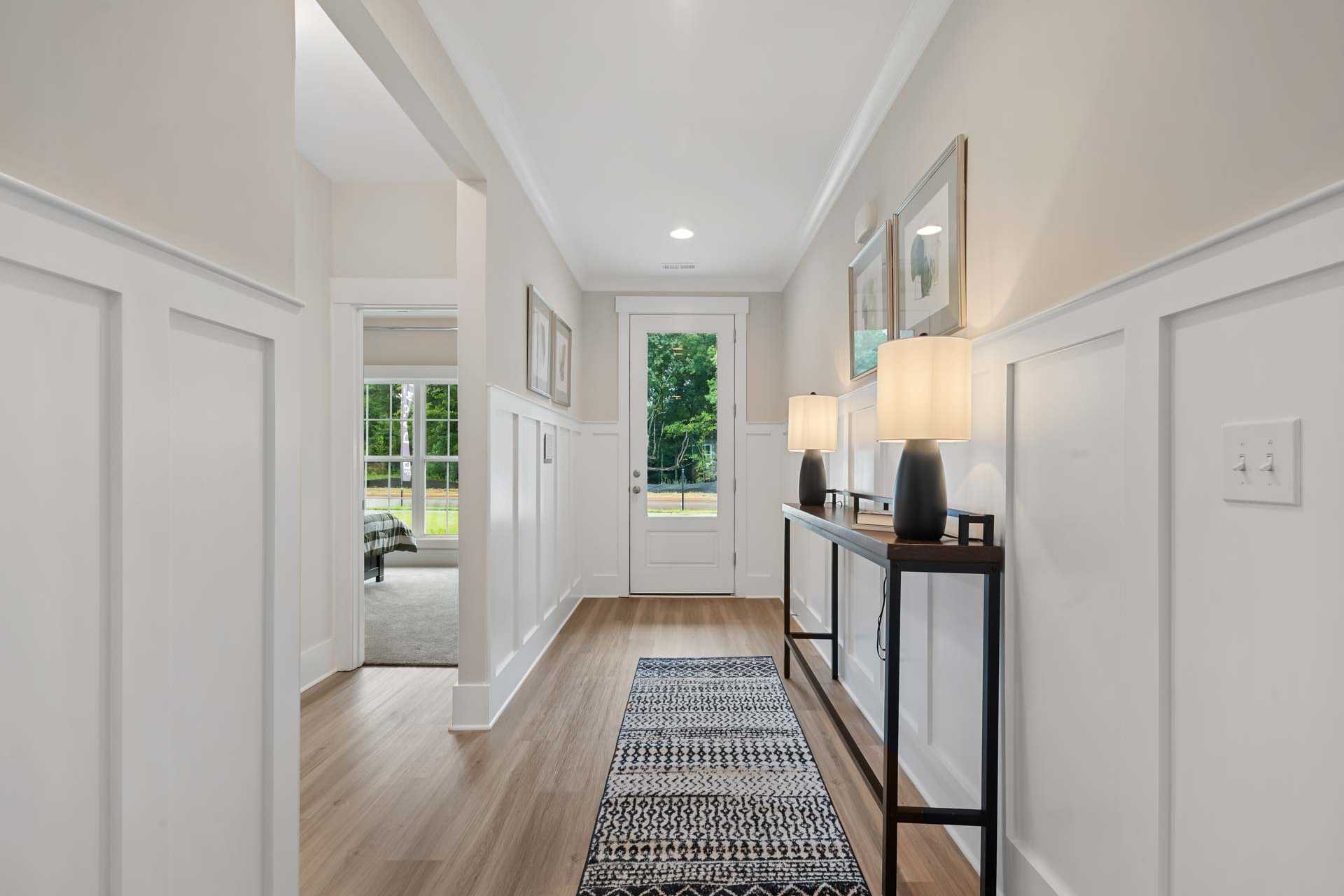 Wainscoted hallway at Durham Farms in Harvest, AL with oak hardwood floors, patterned runner rug, console table lamps, and French doors to greenery