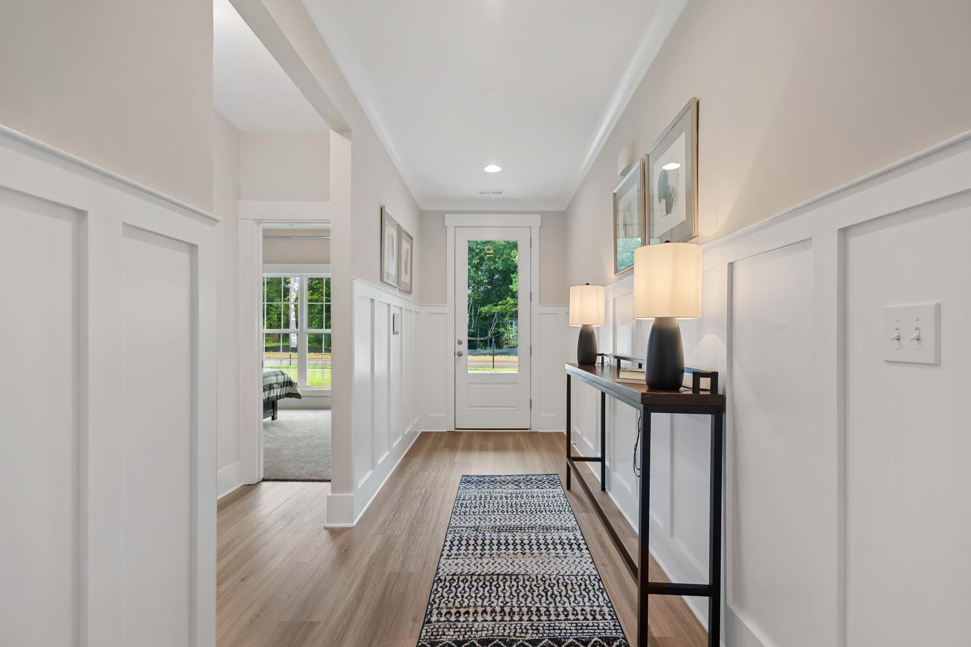 Wainscoted hallway at Durham Farms in Harvest, AL with oak hardwood floors, patterned runner rug, console table lamps, and French doors to greenery