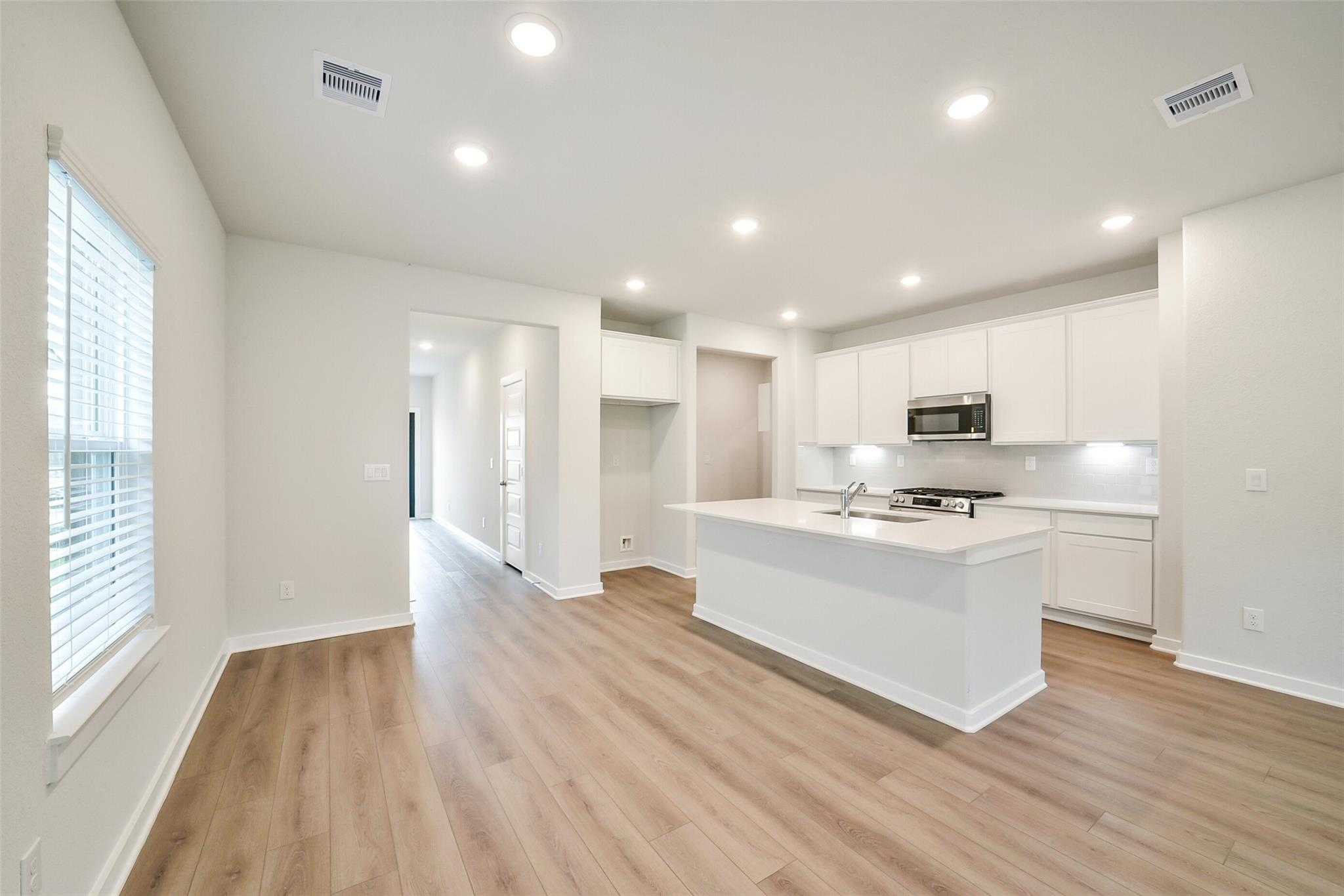 Bright open-concept kitchen with white cabinets, large island sink, stainless appliances, and hardwood floors in The Blanco E, Magnolia, Texas