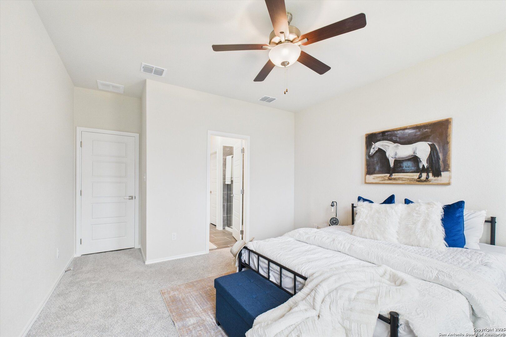 Master bedroom with king bed, white duvet, blue accents, ceiling fan, and en-suite door in Davidson Homes The Asheville K, Bricewood, San Antonio