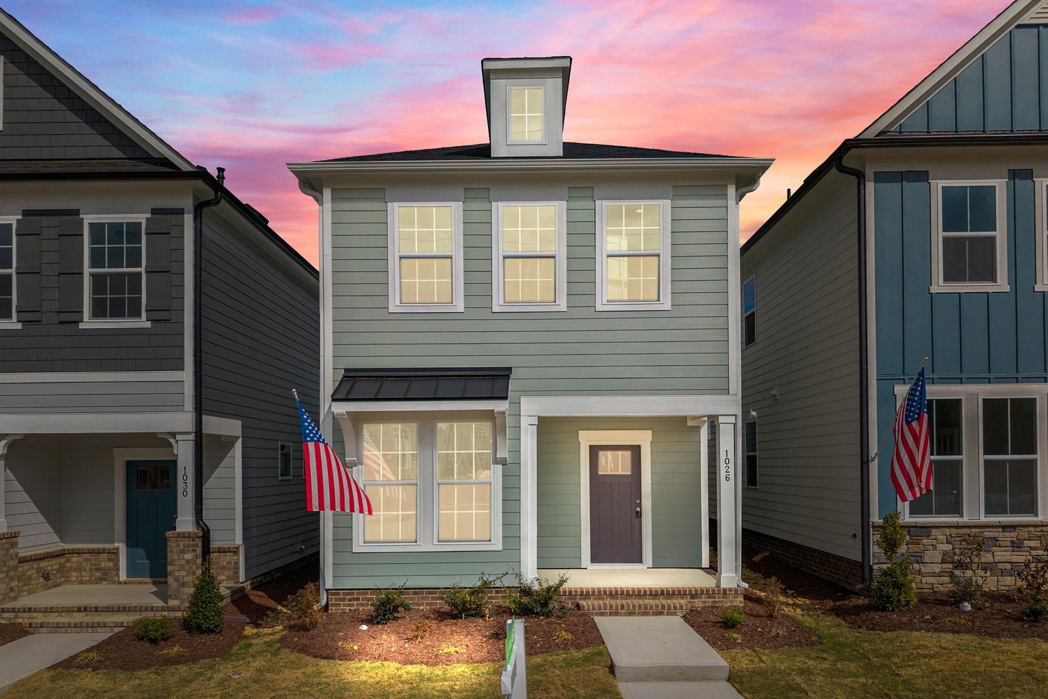 Charming light green Craftsman home exterior at Forestville Yard in Knightdale NC with covered porch, American flag, and sunset sky