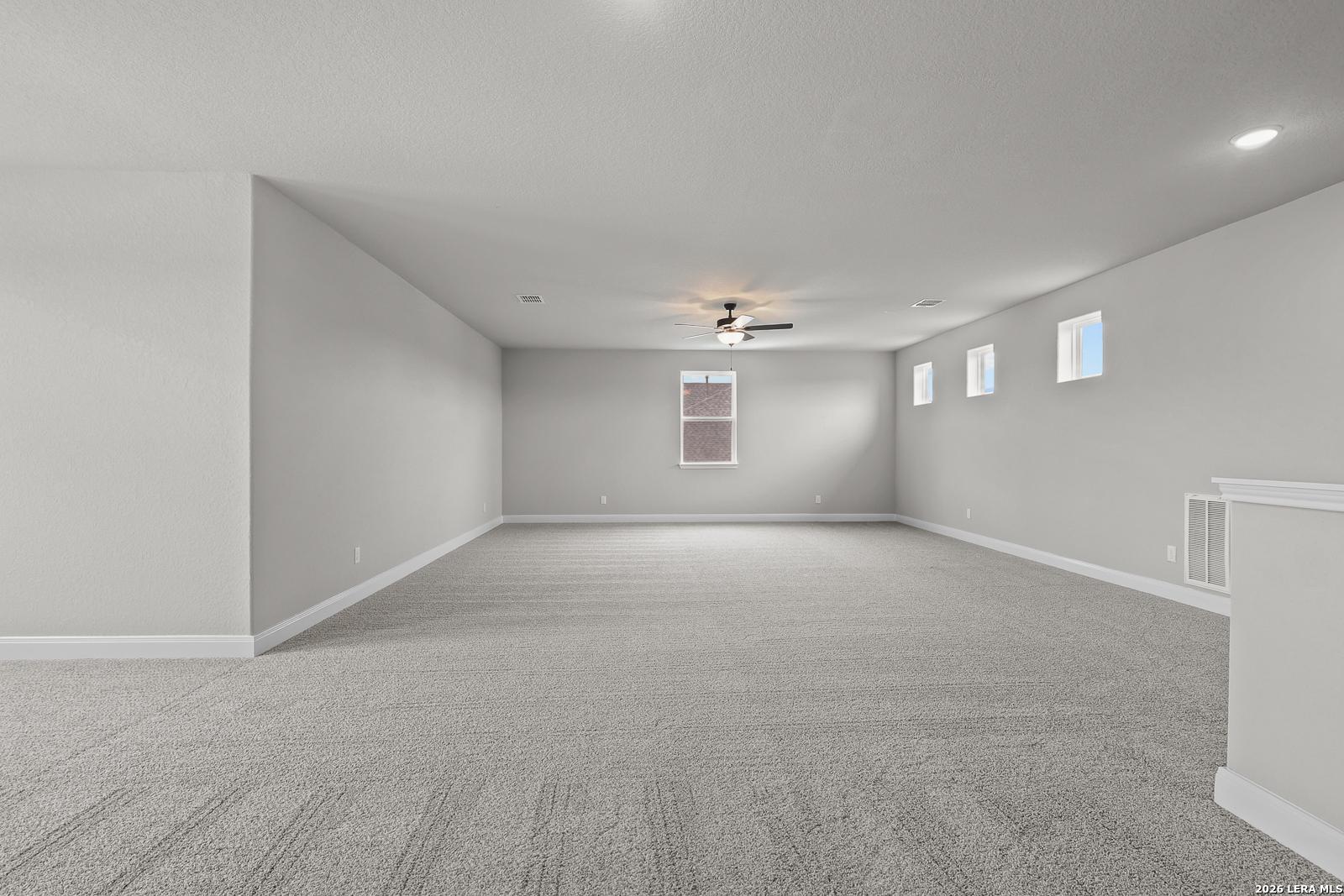 Spacious empty upstairs bonus room with gray carpet, light walls, ceiling fan, recessed lights, and windows in The Belmont H home, Castroville, Texas